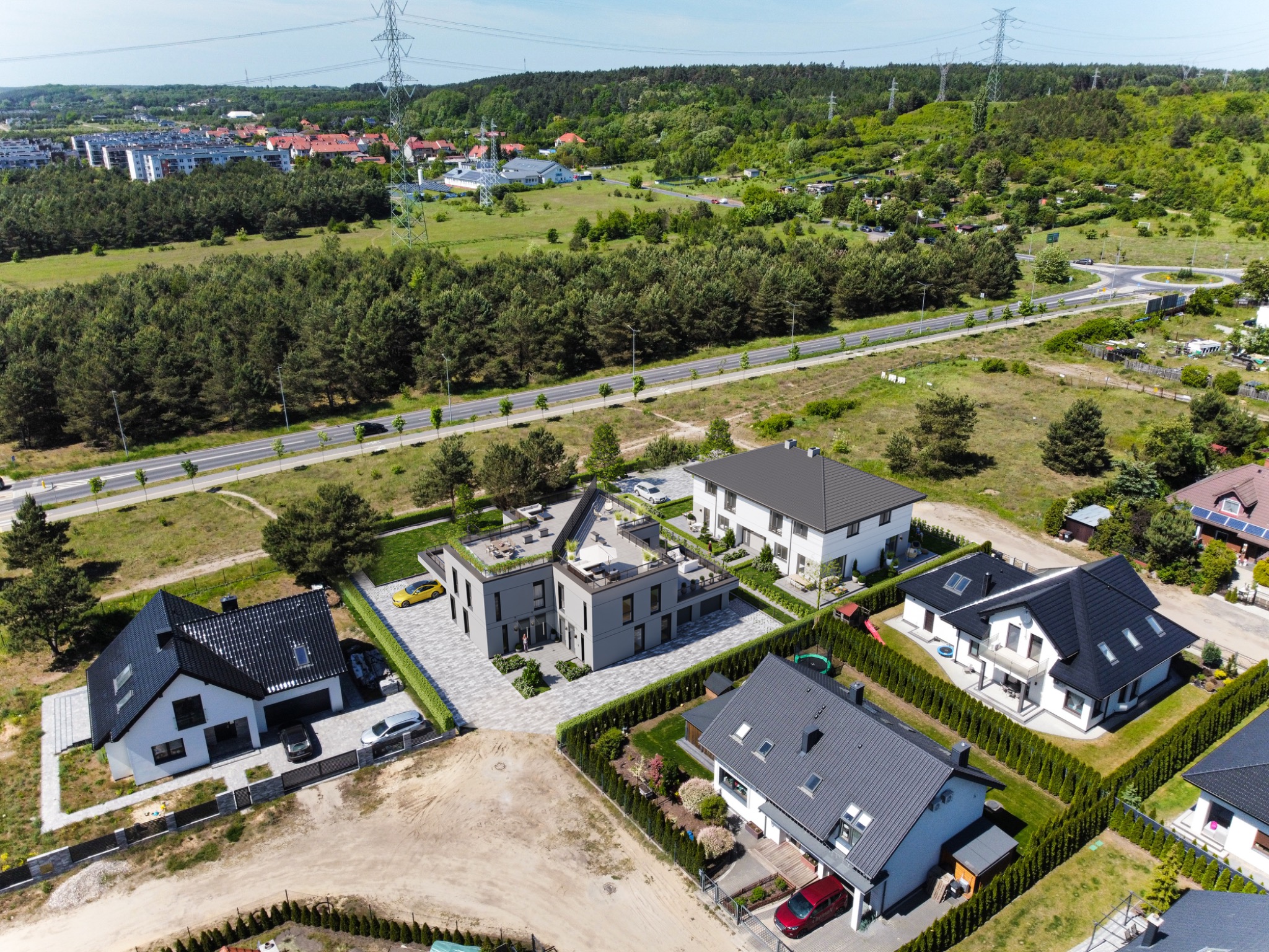 Widok z lotu ptaka na kilka nowoczesnych domów jednorodzinnych z zadbanymi ogrodami, usytuowanych w sąsiedztwie lasu i osiedla bloków, w słoneczny dzień.