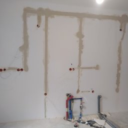 Bohdan Bohdan - Ściana w trakcie prac instalacyjnych: widoczne bruzdy zalane gipsem, wystające przewody elektryczne z puszek oraz instalacja wodna z rurami w kolorach niebieskim i czerwonym.