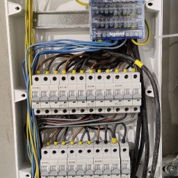 Bohdan Bohdan - Otwarta skrzynka bezpiecznikowa z zainstalowanymi bezpiecznikami Eaton, listwami zaciskowymi i przewodami o różnych kolorach, zamontowana na szarej, surowej ścianie.