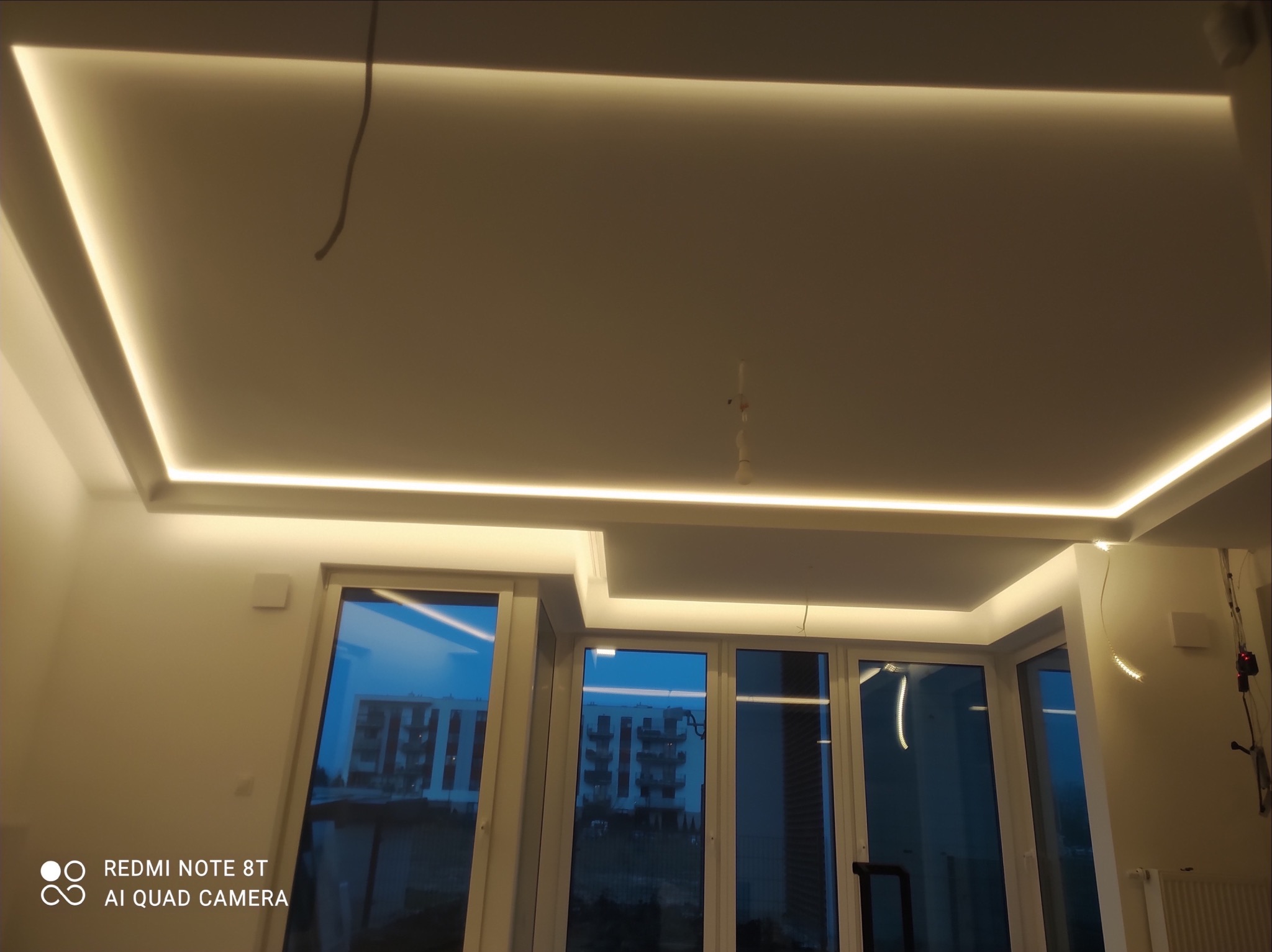 Nowoczesny sufit podwieszany z oświetleniem LED w kształcie prostokąta, widoczne okna z odbiciem zabudowy miejskiej.