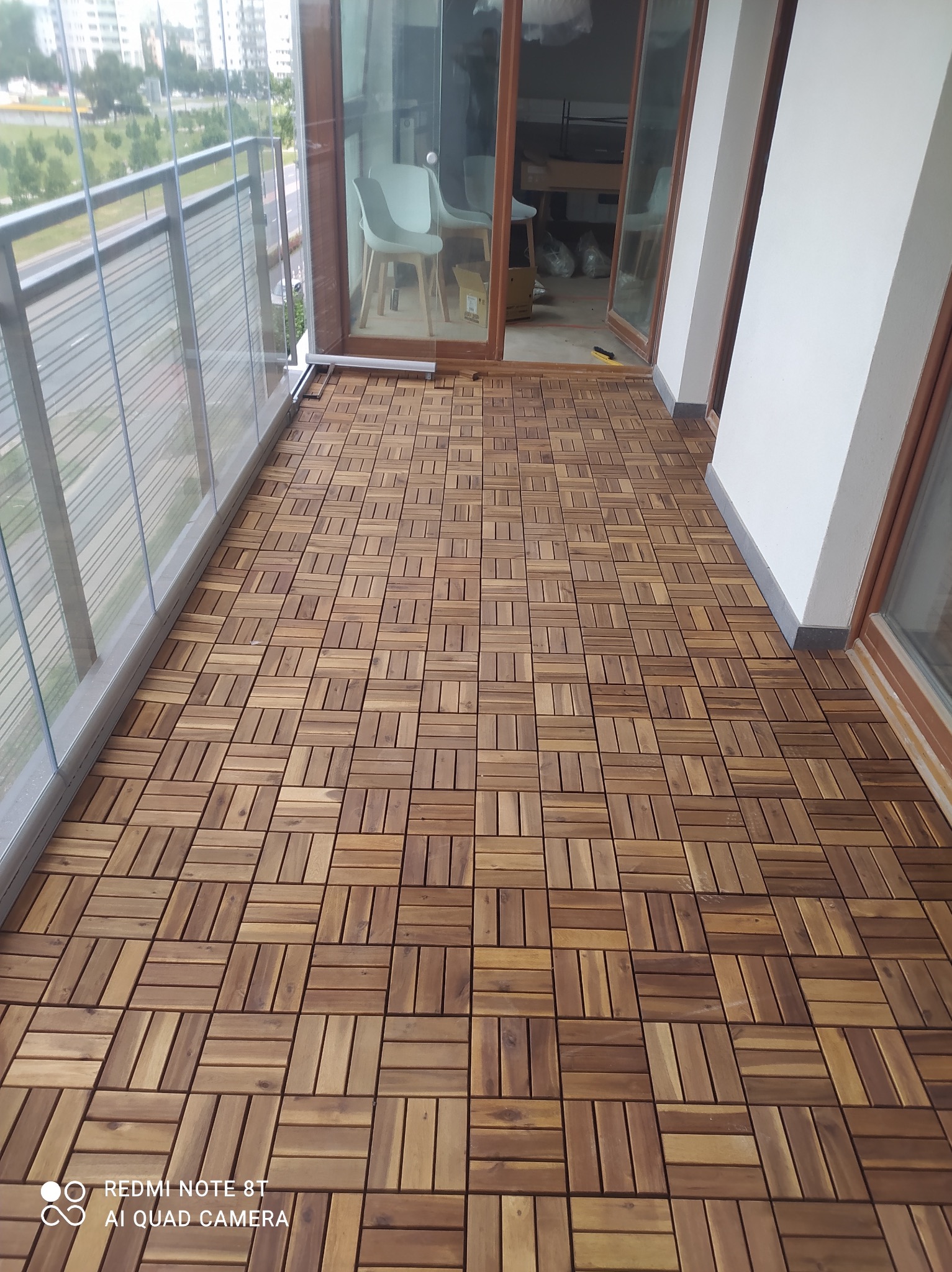 Wykończony balkon z drewnianą podłogą z kwadratowych płytek, szklana balustrada, widok na zewnątrz.