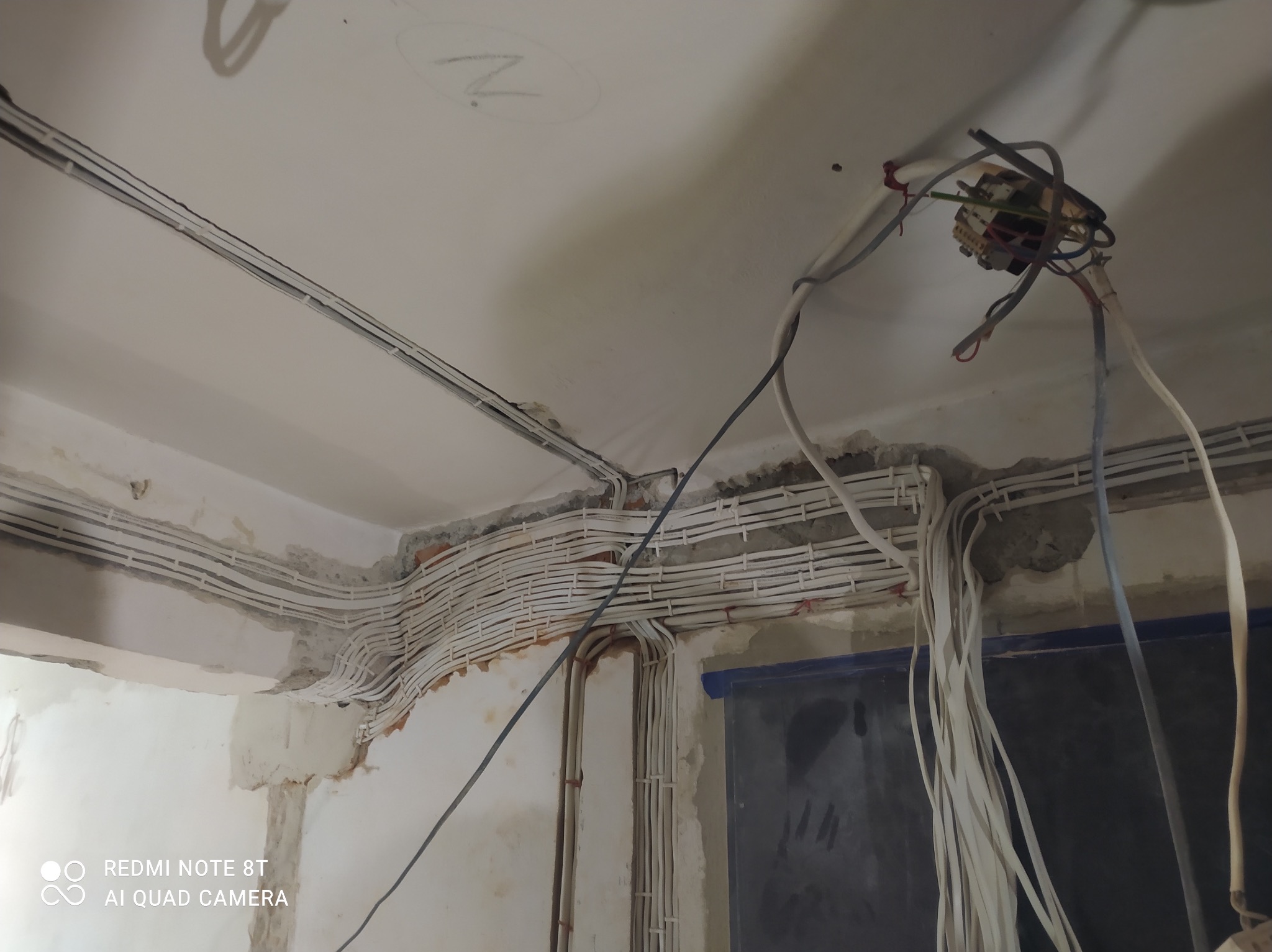 Instalacja elektryczna w trakcie montażu: kable ułożone w korytkach na ścianie pod sufitem, widoczna puszka elektryczna z wystającymi przewodami, ściany w stanie surowym z widocznymi śladami po kuciu.
