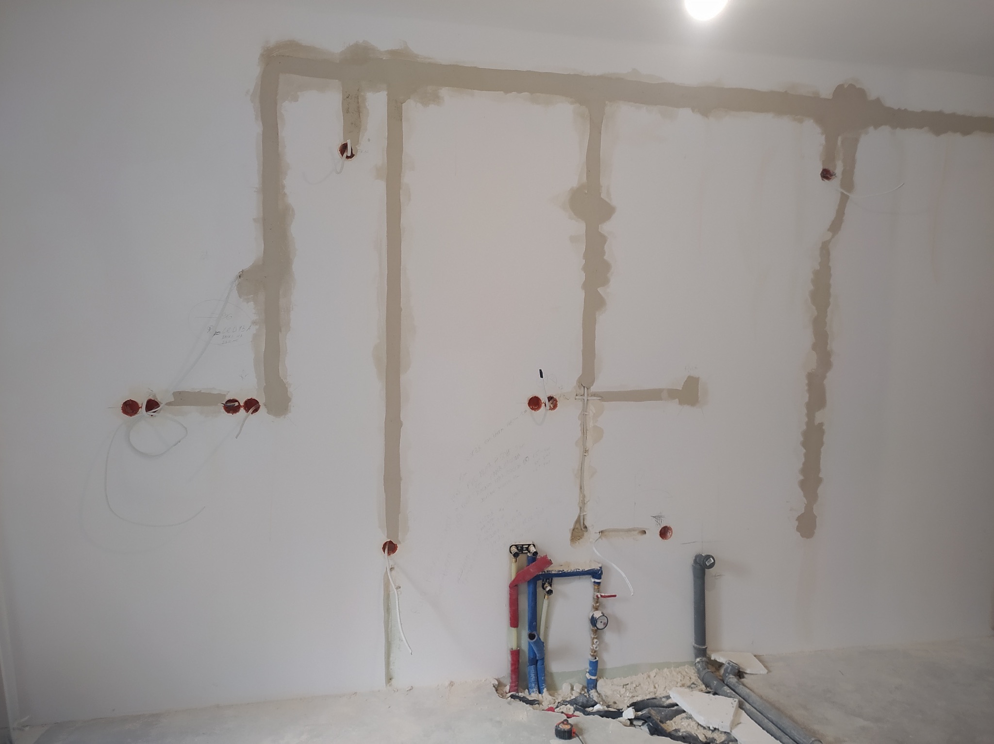 Ściana w trakcie prac instalacyjnych: widoczne bruzdy zalane gipsem, wystające przewody elektryczne z puszek oraz instalacja wodna z rurami w kolorach niebieskim i czerwonym.