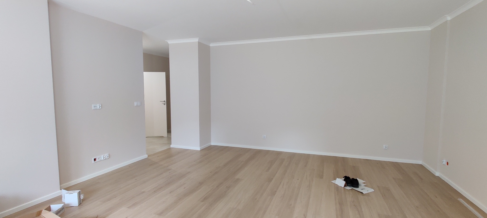 Pomalowane na beżowo puste pomieszczenie z drewnianą podłogą, białymi listwami przypodłogowymi i sztukaterią sufitową; widoczne gniazdka elektryczne, otwarte drzwi do sąsiedniego pomieszczenia...