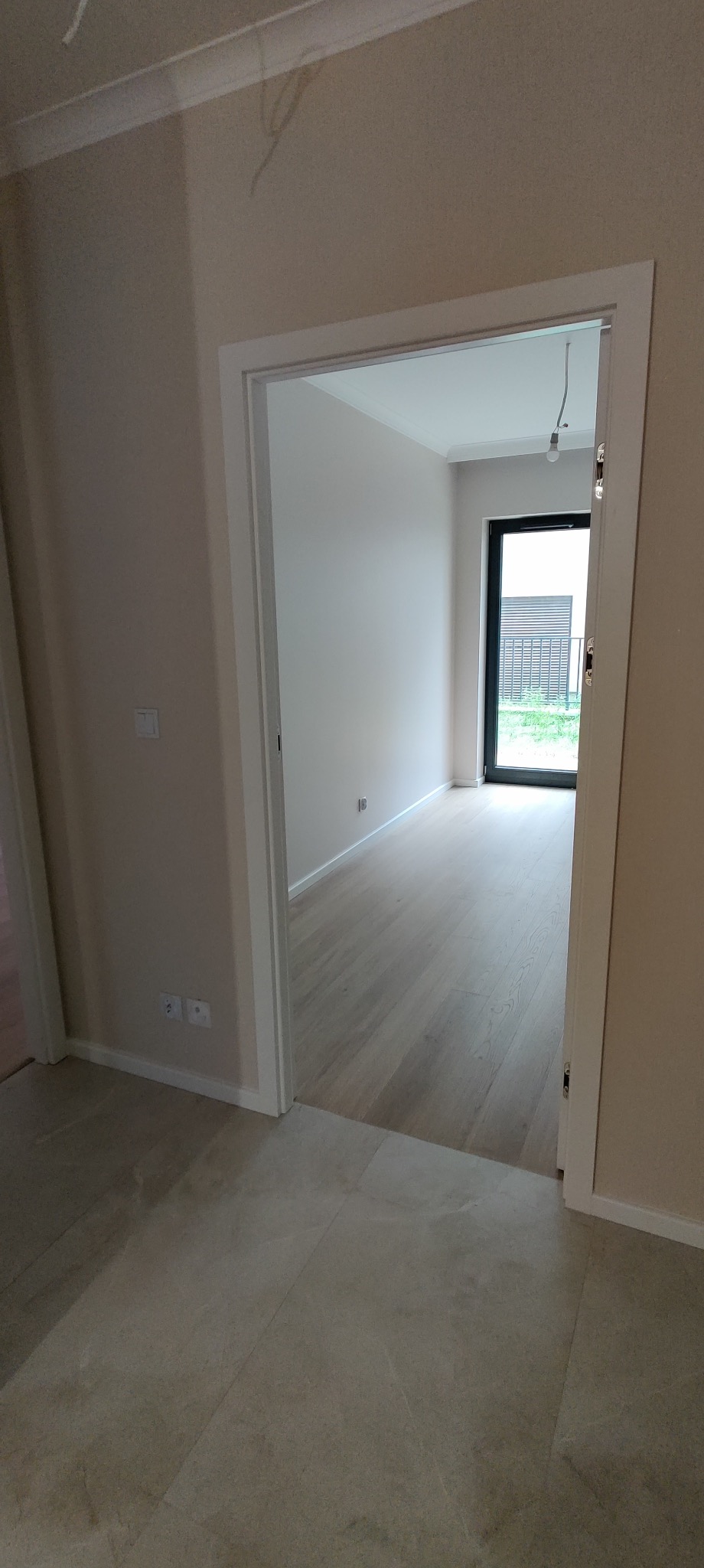 Widok przez białe futryny drzwi na jasny pokój z drewnianą podłogą i czarnymi drzwiami balkonowymi, widoczny fragment podłogi z płytek.