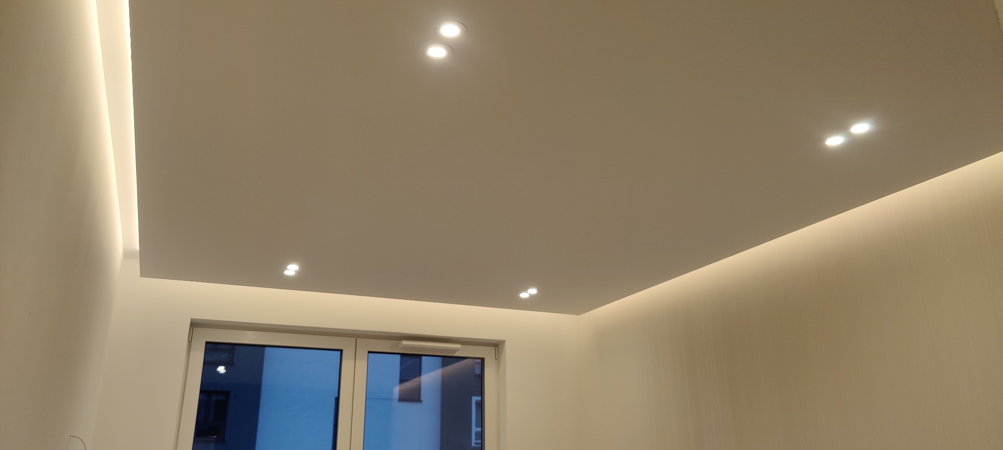 Podwieszany sufit z oświetleniem LED wzdłuż krawędzi i punktowymi lampami wpuszczanymi, widok na okno z odbiciem.