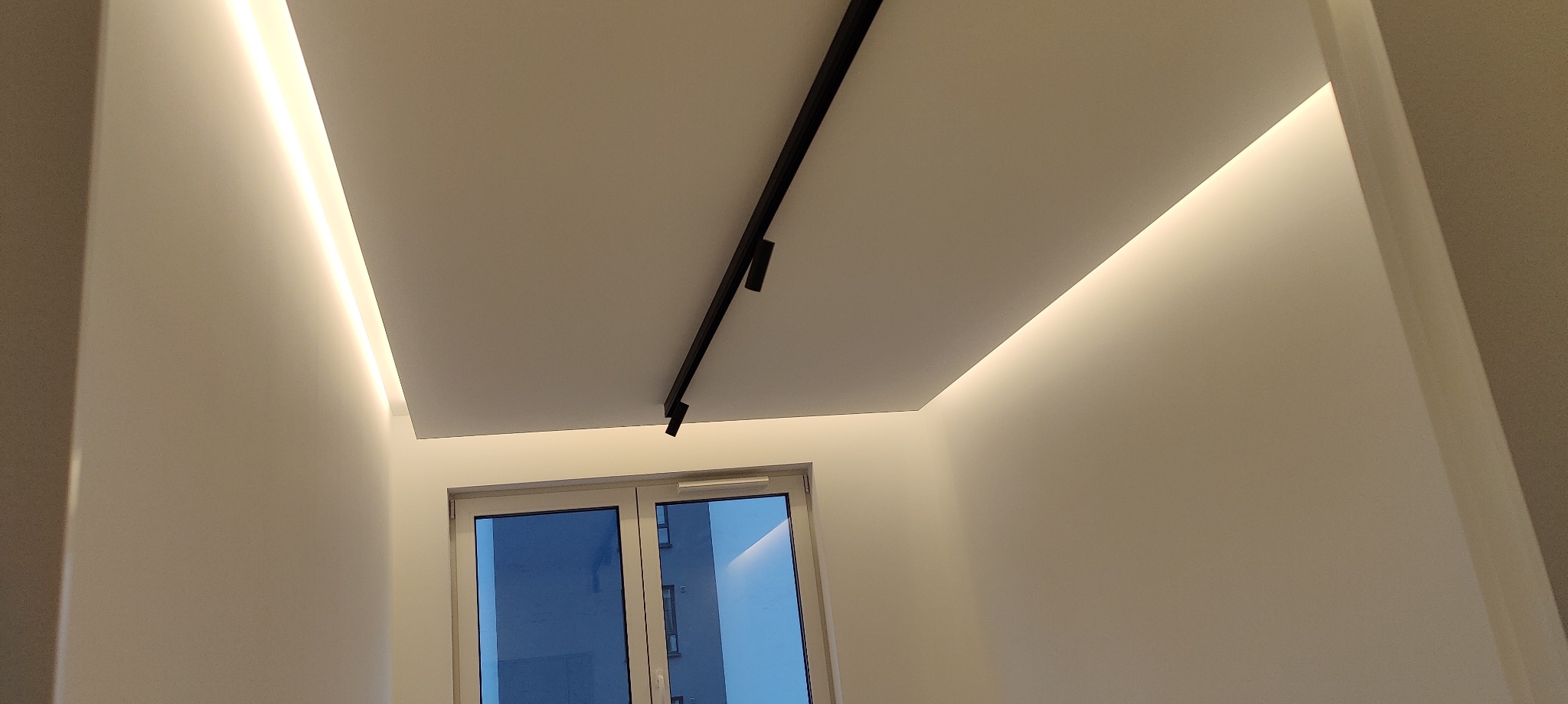 Nowoczesne oświetlenie sufitowe z szynoprzewodem i ukrytymi listwami LED w białym wnętrzu z oknem.