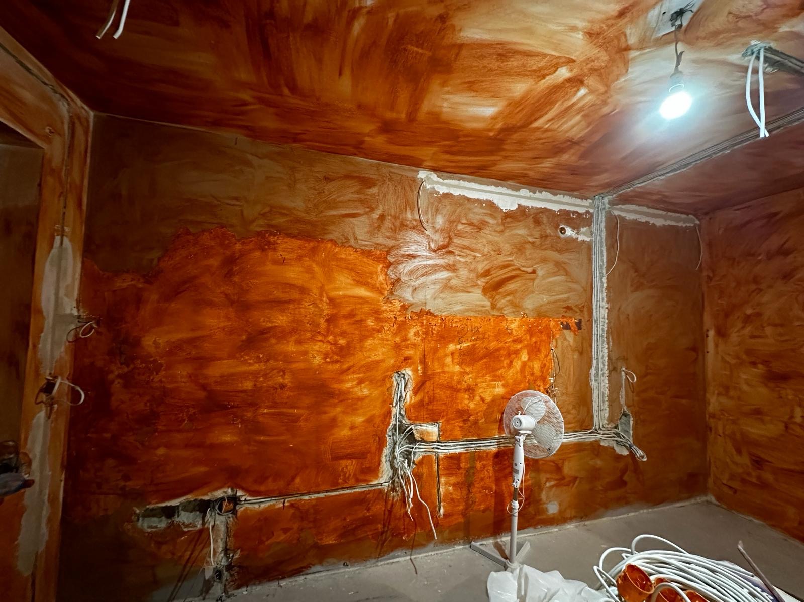 Pokój w trakcie prac przygotowawczych do malowania, z nałożoną warstwą gruntującą o brązowym odcieniu, widoczne instalacje elektryczne i wentylator stojący.