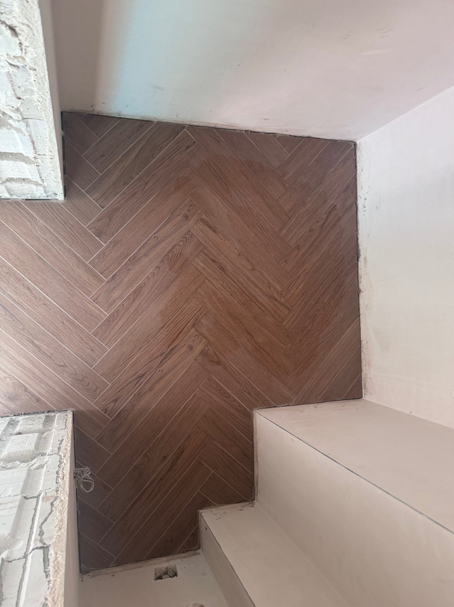 Ułożona brązowa podłoga z płytek imitujących drewno w jodełkę, widoczny fragment schodów i surowa ściana z przewodami.