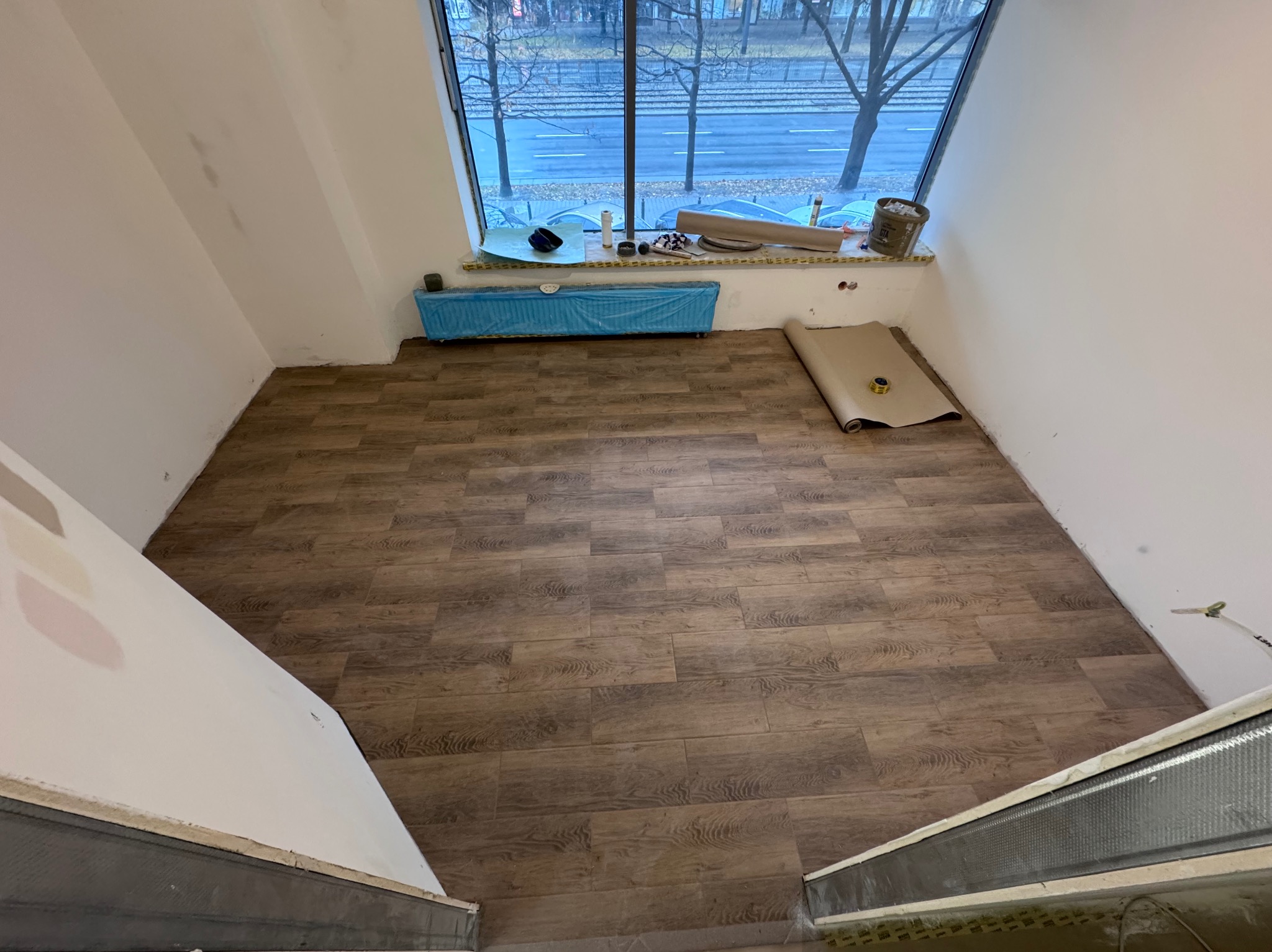Wnętrze pokoju w trakcie remontu, z nową podłogą imitującą drewno, okno z widokiem na ulicę, zabezpieczony grzejnik i przygotowane materiały do malowania.