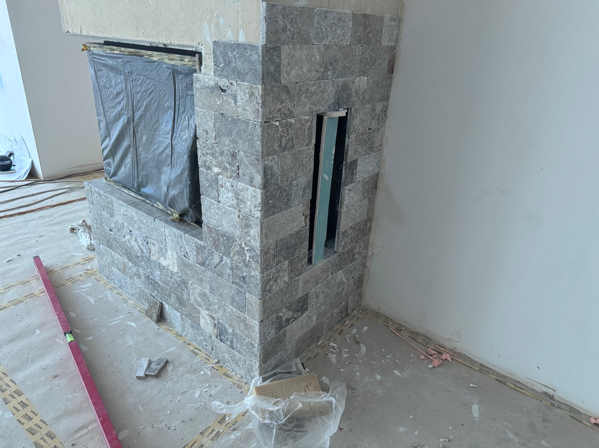 Obróbka kominka z szarego kamienia bloczkowego, zabezpieczone palenisko folią, poziomica i resztki materiałów na podłodze.