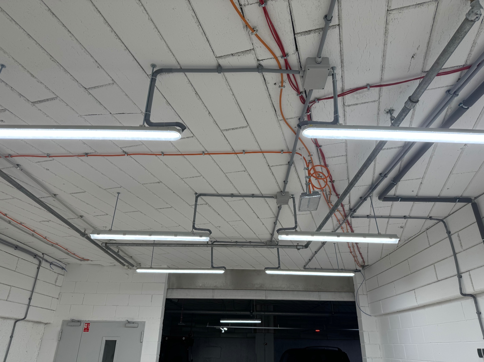 Sufit z widoczną instalacją elektryczną: rurki PCV, kable w kolorach czerwonym i pomarańczowym, puszki elektryczne i lampy jarzeniowe, surowy mur z białej cegły