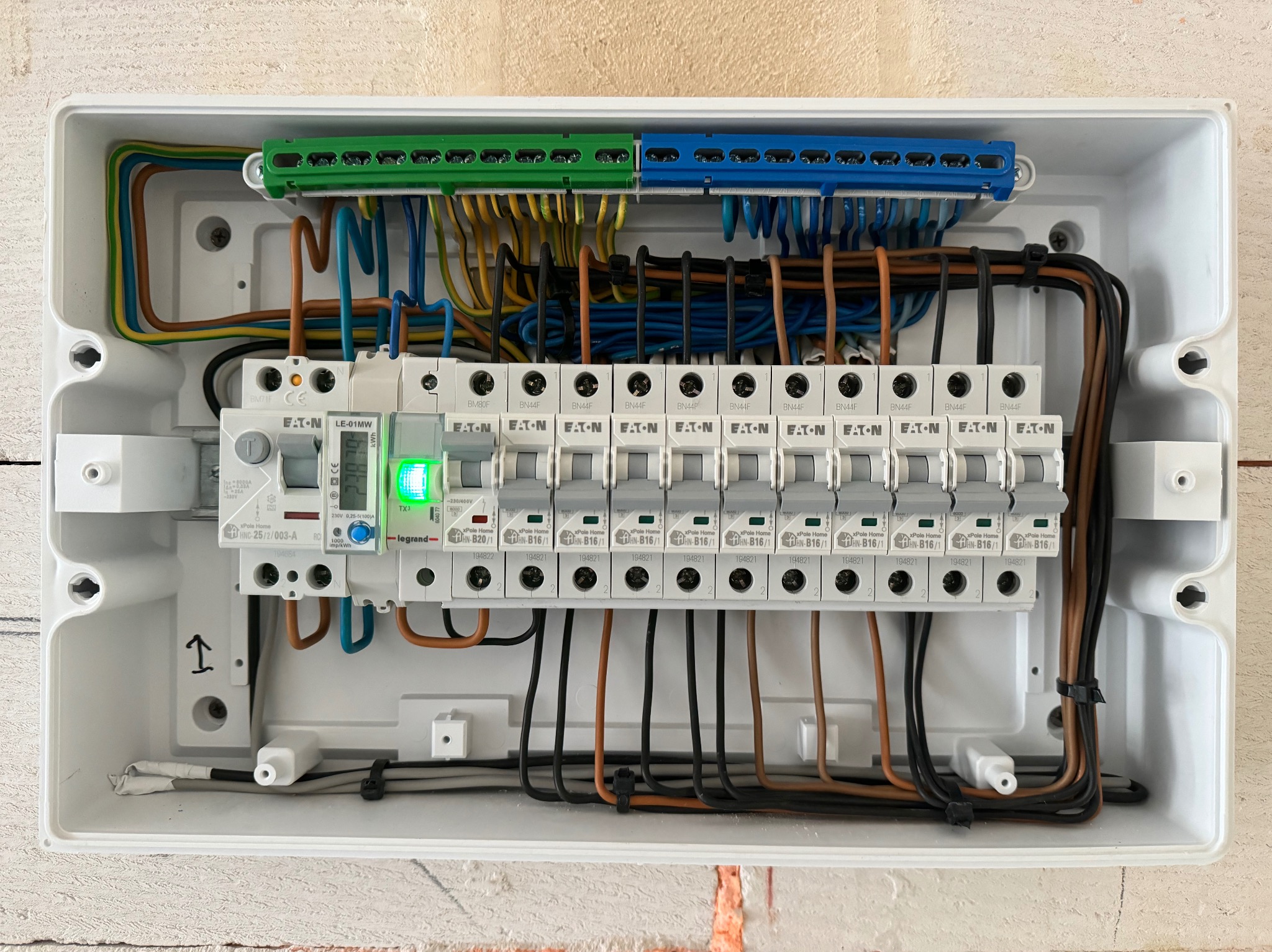 Otwarta skrzynka elektryczna z zainstalowanymi bezpiecznikami EATON, licznikiem Legrand i uporządkowanymi przewodami w kolorach brązowym, niebieskim, żółto-zielonym i czarnym.