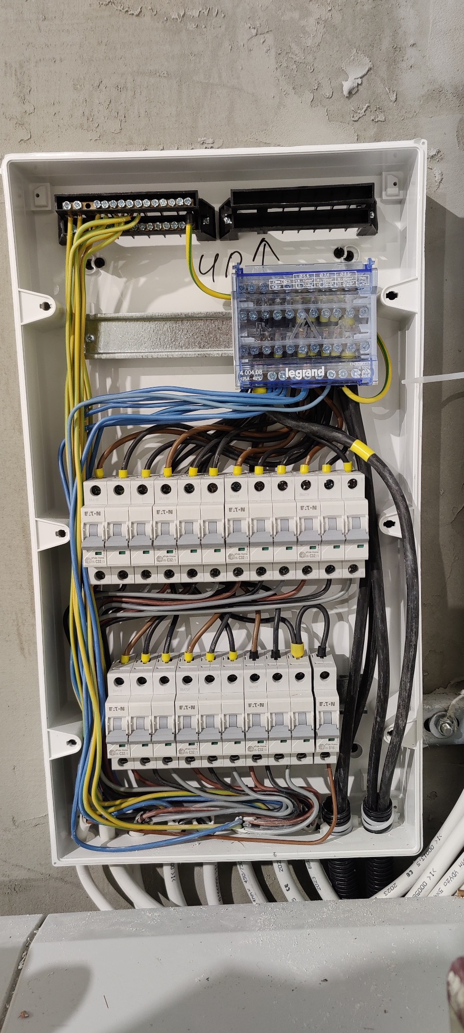 Otwarta skrzynka bezpiecznikowa z zainstalowanymi bezpiecznikami Eaton, listwami zaciskowymi i przewodami o różnych kolorach, zamontowana na szarej, surowej ścianie.