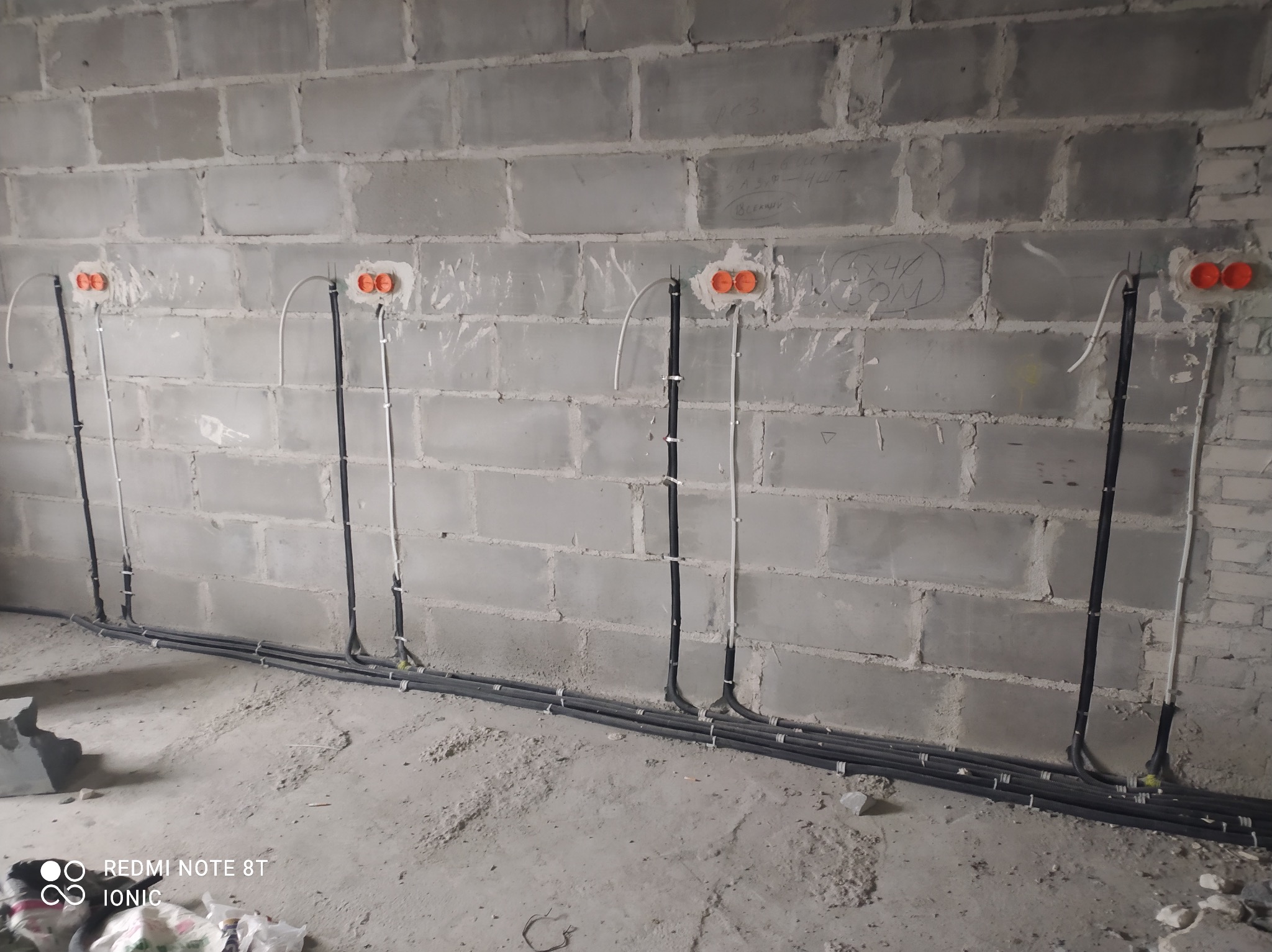 Surowy mur z betonu komórkowego z widocznymi przewodami elektrycznymi w peszlach, prowadzącymi do pomarańczowych puszek instalacyjnych.