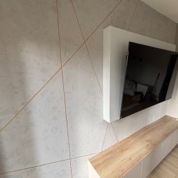 GRAB-TECH Aleksandra Grabowska - Ściana z dekoracyjną fakturą i wstawkami w kolorze miedzi, podwieszony telewizor z białą obudową i szafka RTV z drewnianym blatem, parkiet na podłodze.