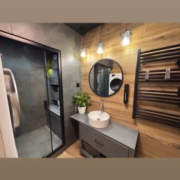 GRAB-TECH Aleksandra Grabowska - Nowoczesna łazienka z kabiną prysznicową typu walk-in, okrągłym lustrem odbijającym pralkę, umywalką nablatową na szarej szafce, grzejnikiem drabinkowym i drewnopodobnymi płytkami na ścianie.