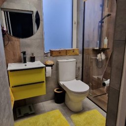 GRAB-TECH Aleksandra Grabowska - Nowoczesna łazienka z szarymi płytkami, żółtymi akcentami w postaci szafki pod umywalkę i dywaników, okrągłe lustro z czarną ramą i kabina prysznicowa z drewnopodobnymi panelami.