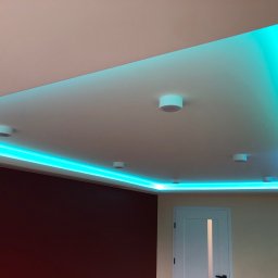 GRAB-TECH Aleksandra Grabowska - Podwieszany sufit z oświetleniem LED w kolorze turkusowym, uzupełniony punktowymi lampami sufitowymi, widok na fragment ściany w kolorze burgundowym oraz białe drzwi.