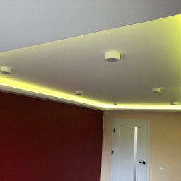 GRAB-TECH Aleksandra Grabowska - Podwieszany sufit z oświetleniem LED wzdłuż krawędzi, uzupełniony okrągłymi lampami sufitowymi, na tle ścian w odcieniach bordo i beżu oraz białych drzwi.