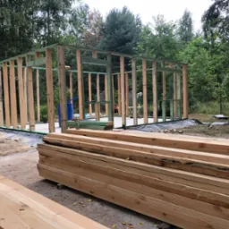 Konstrukcja szkieletowa budynku w trakcie budowy, widoczne drewniane belki i słupy, stos drewna na pierwszym planie, teren budowy zalesiony w tle.