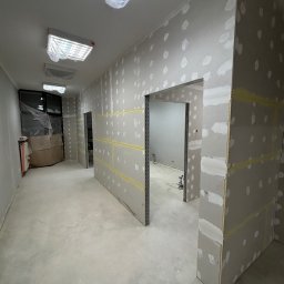 GRAB-TECH Aleksandra Grabowska - Wnętrze w trakcie prac wykończeniowych, widoczne płyty kartonowo-gipsowe z widocznymi punktami szpachlowania, metalowe profile ościeżnic, zabezpieczone folią lampy sufitowe.