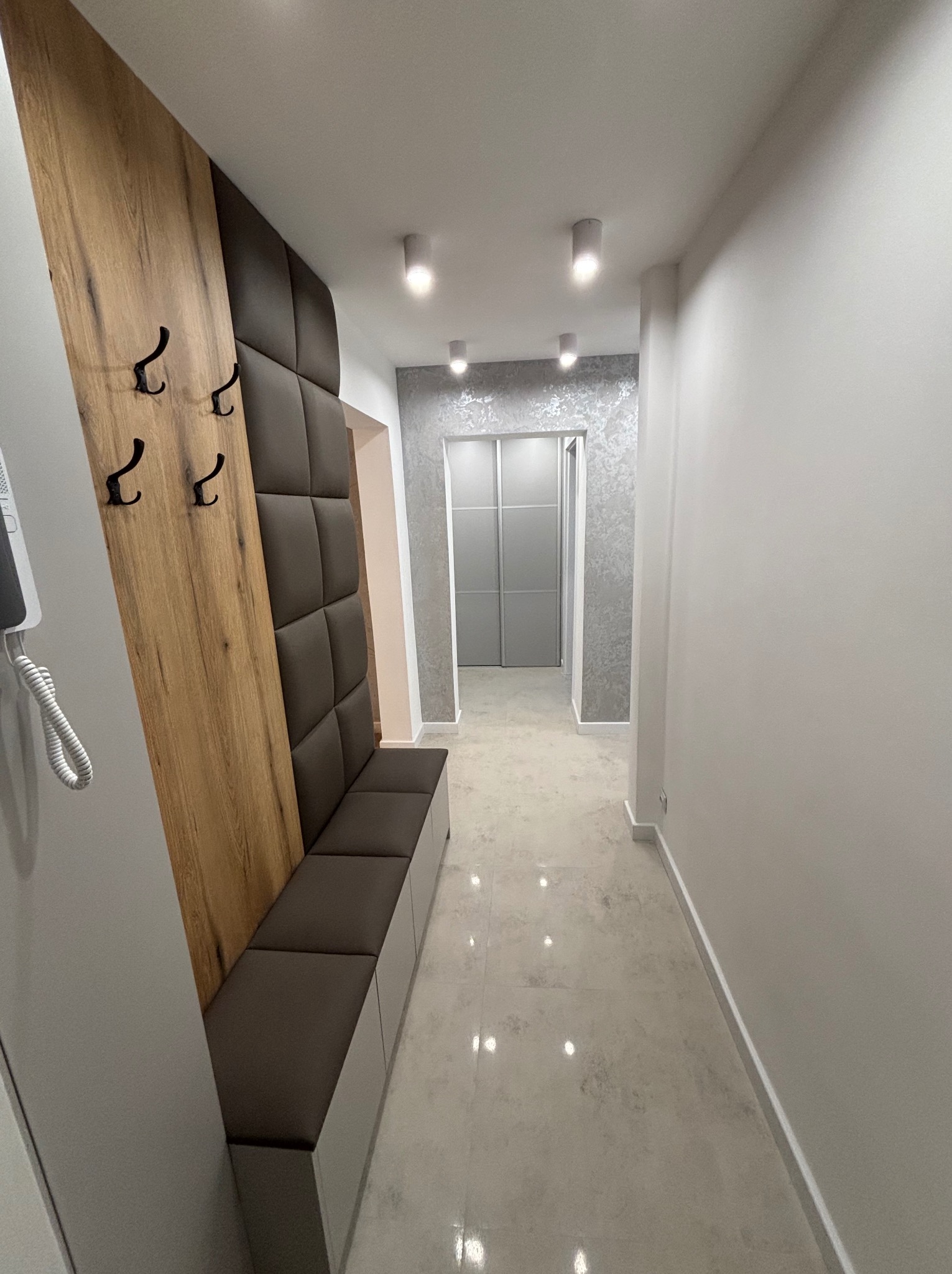 Nowoczesny przedpokój z panelem drewnopodobnym, tapicerowanym siedziskiem w kolorze ciemnego brązu i czarnymi wieszakami, wykończony jasnymi płytkami podłogowymi i srebrną tapetą w głębi.