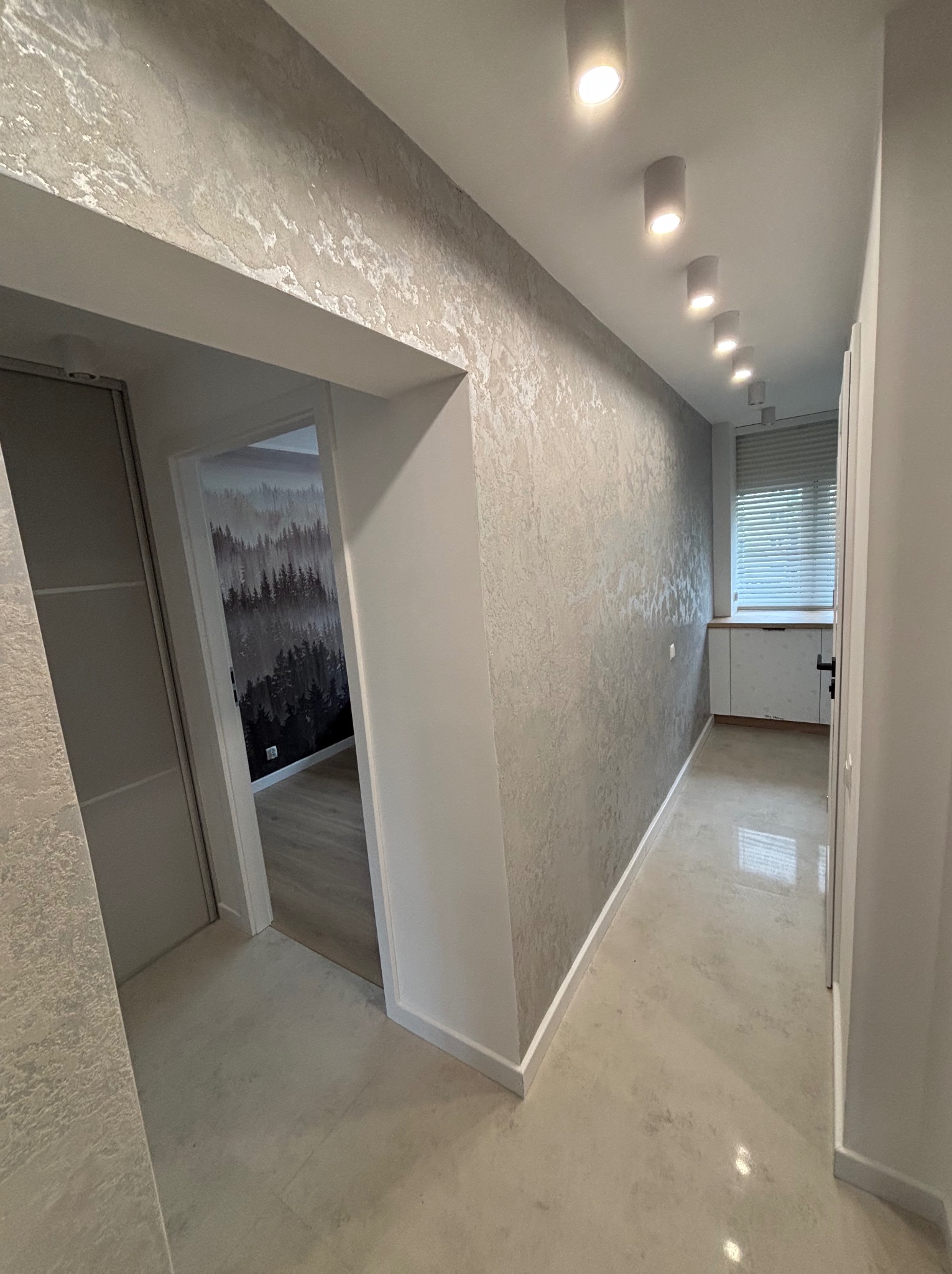 Korytarz po remoncie z dekoracyjną, srebrzystą strukturą na ścianach, punktowe oświetlenie sufitowe i widok na pokój z fototapetą leśną.