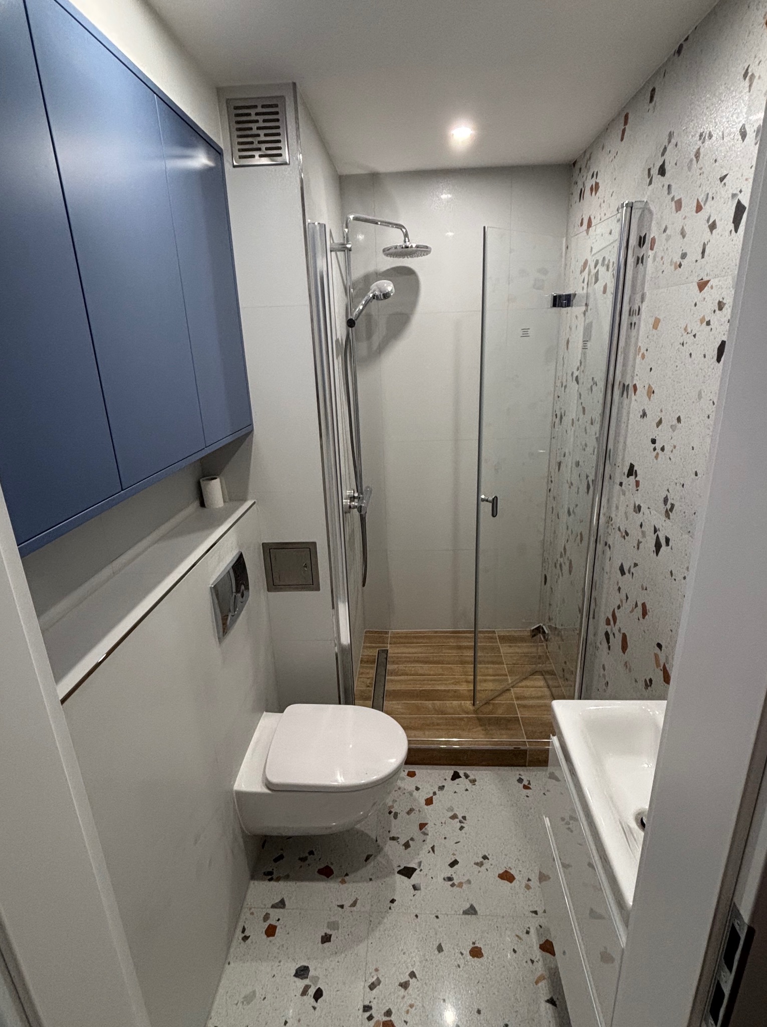 Nowoczesna łazienka z kabiną prysznicową typu walk-in, podwieszaną toaletą i szafką w kolorze granatowym, wykończona płytkami lastryko na podłodze i ścianie akcentowej.