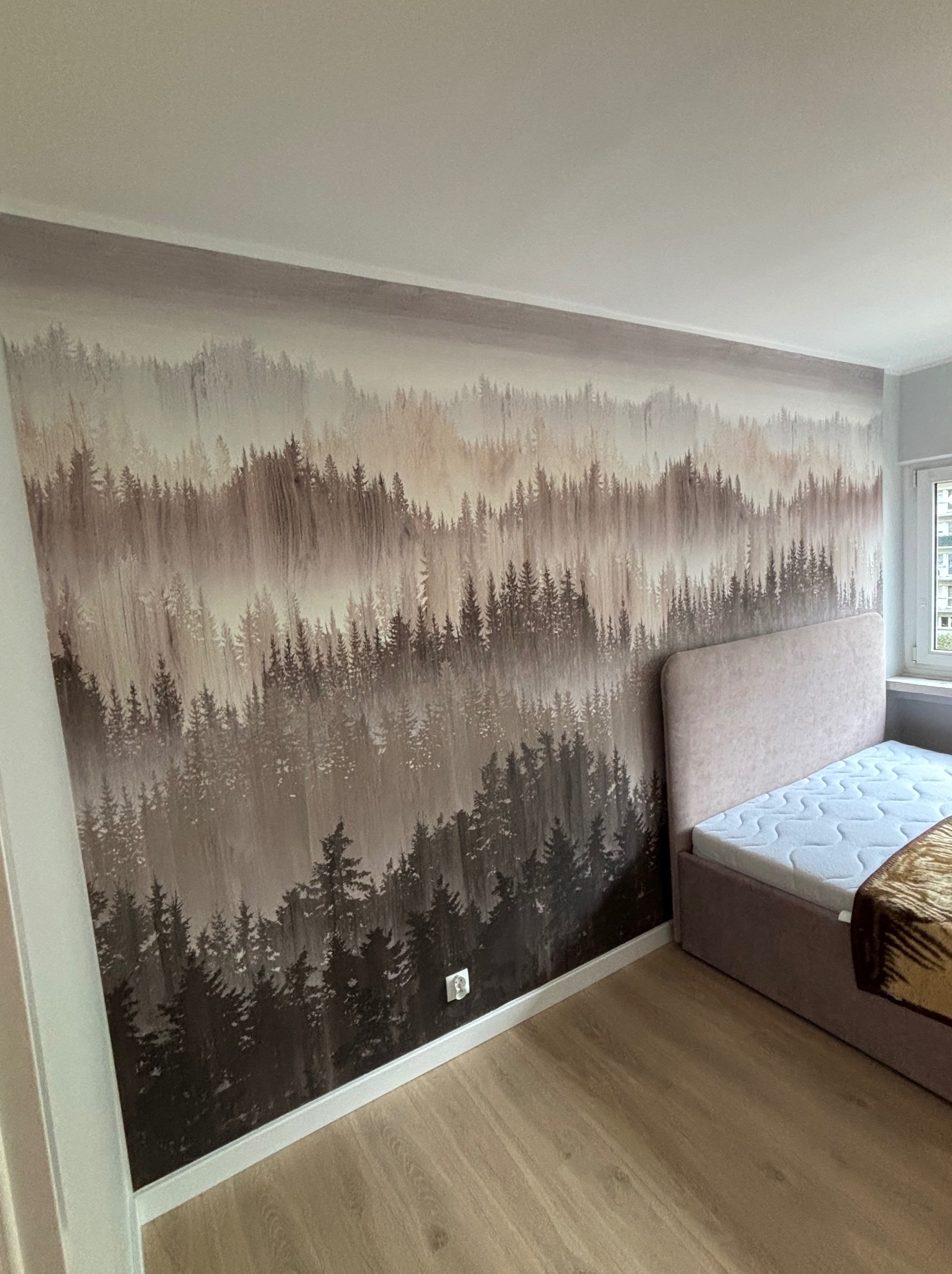 Sypialnia z fototapetą przedstawiającą las we mgle, z łóżkiem z jasnoróżowym zagłówkiem i drewnianą podłogą.