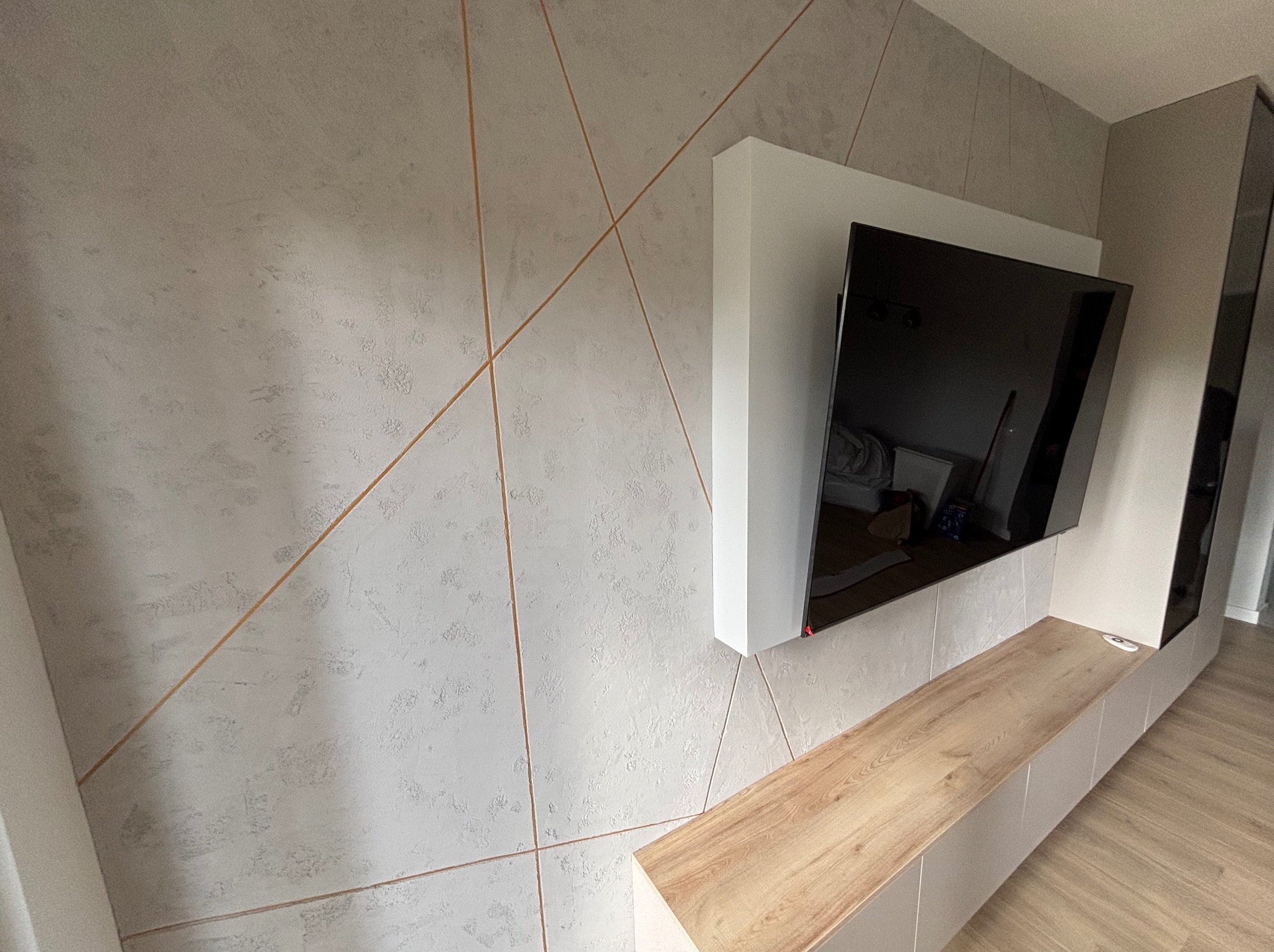 Ściana z dekoracyjną fakturą i wstawkami w kolorze miedzi, podwieszony telewizor z białą obudową i szafka RTV z drewnianym blatem, parkiet na podłodze.