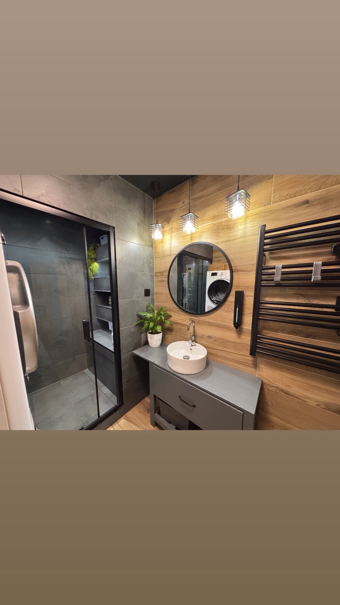 Nowoczesna łazienka z kabiną prysznicową typu walk-in, okrągłym lustrem odbijającym pralkę, umywalką nablatową na szarej szafce, grzejnikiem drabinkowym i drewnopodobnymi płytkami na ścianie.
