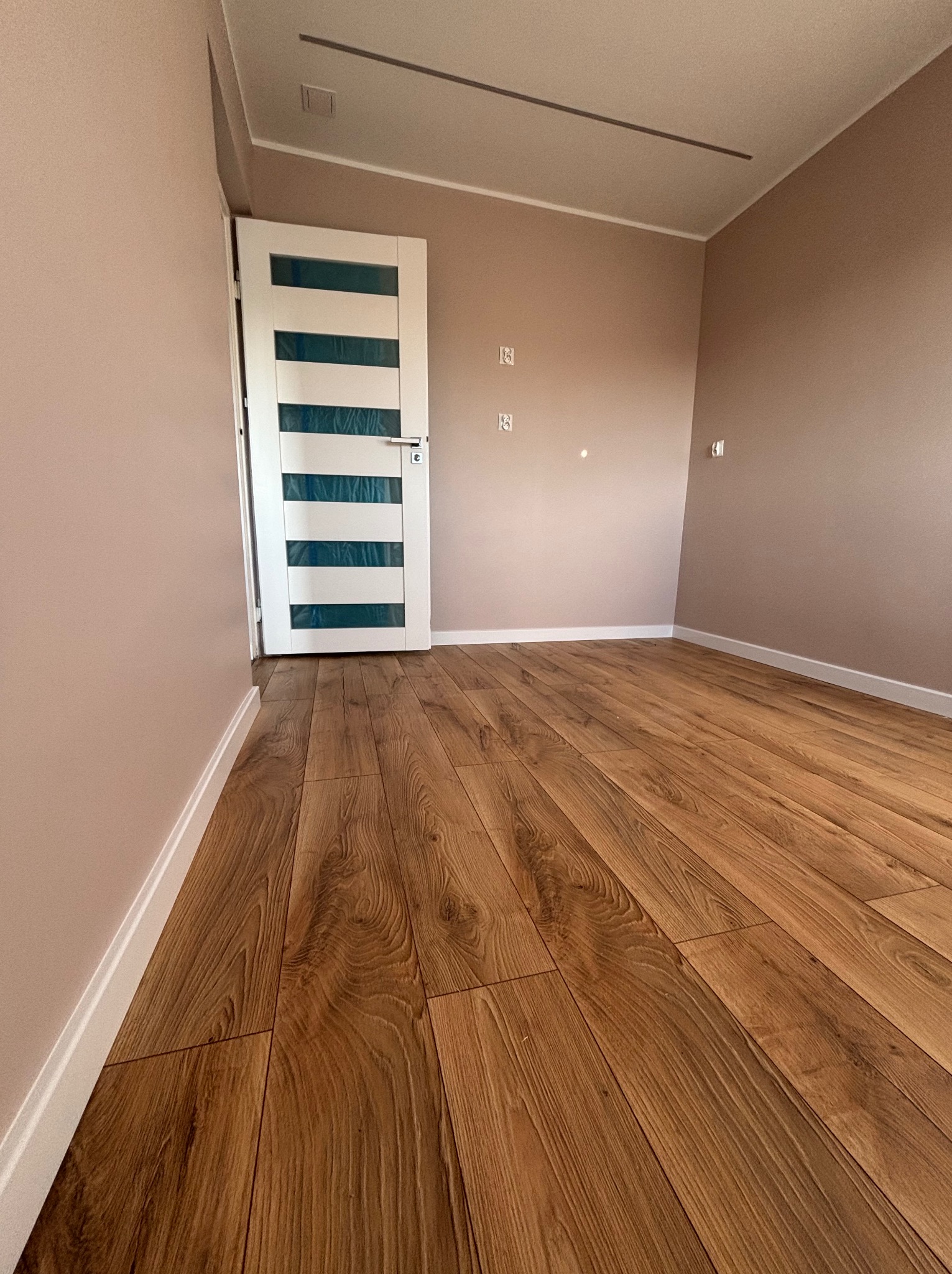 Ukończone wnętrze pokoju z drewnianą podłogą z paneli, pomalowanymi na beżowo ścianami i białą, nowoczesną stolarką drzwiową z przeszkleniami w paski.