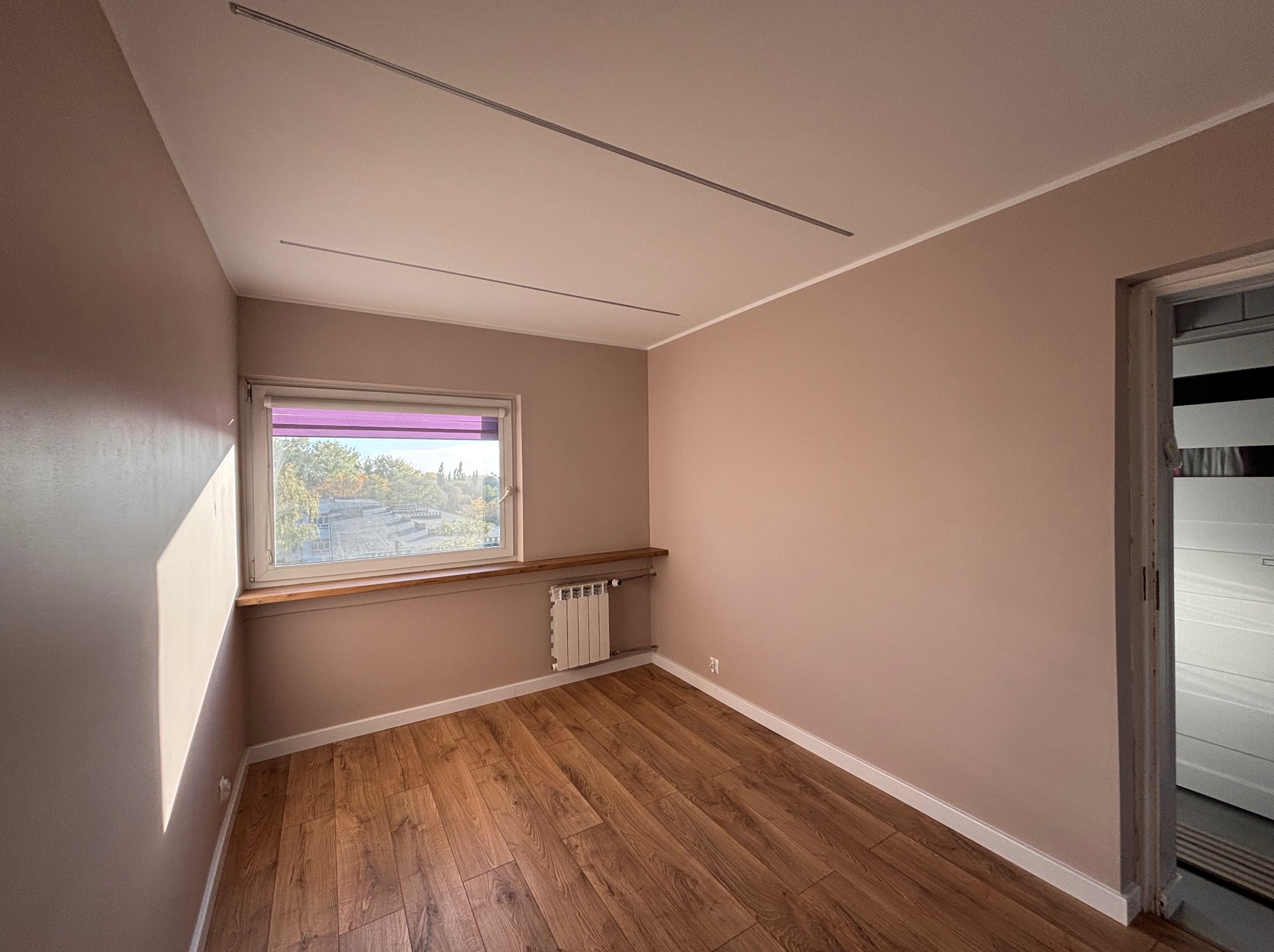 Pokój po remoncie: ściany pomalowane na jasny, ciepły odcień różu, drewniana podłoga, biały grzejnik pod oknem z drewnianym parapetem, widoczne listwy przypodłogowe i szyny oświetleniowe na suficie.