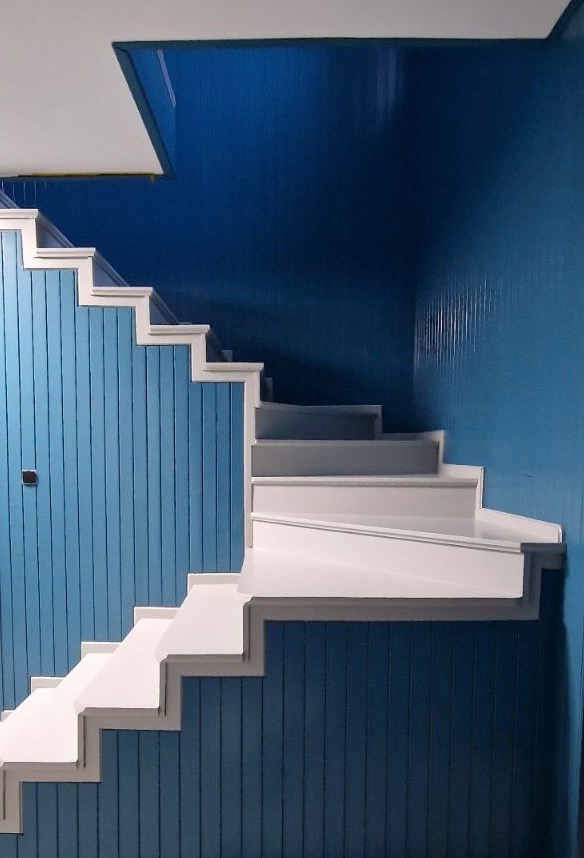 Schody z białymi stopniami i błękitną boazerią ścienną, widok z dołu, pionowe panele na ścianie, kontrastowe kolory.