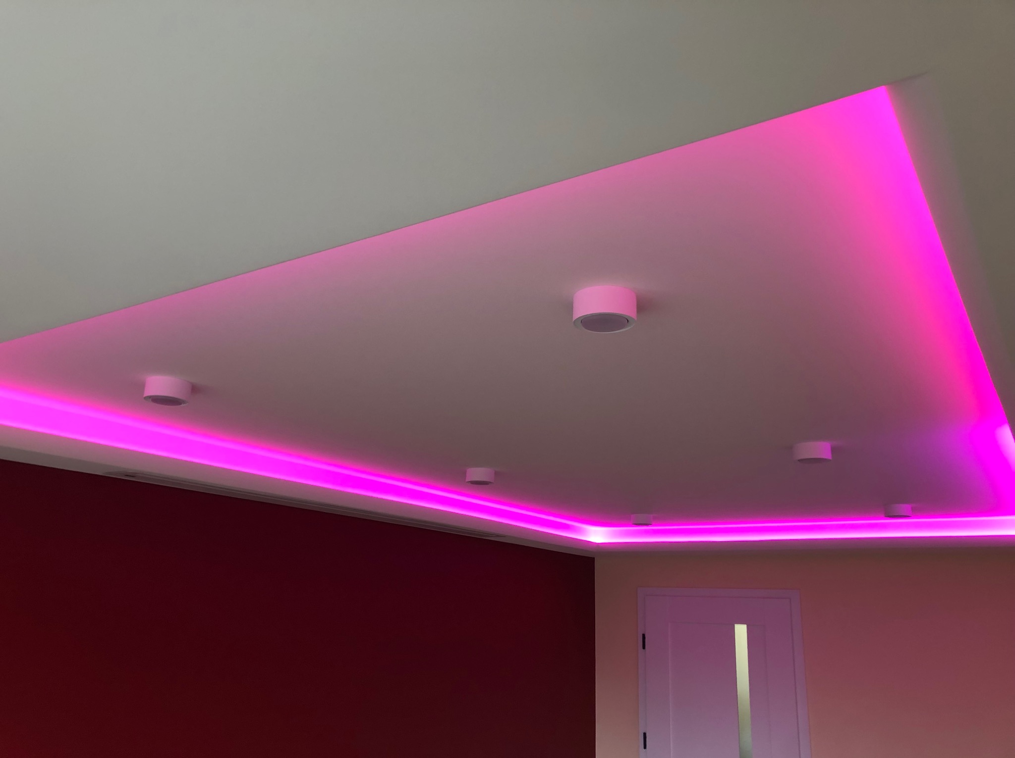 Sufit podwieszany z oświetleniem LED w kolorze różowym, z widocznymi punktowymi lampami, na tle bordowej ściany i białych drzwi.