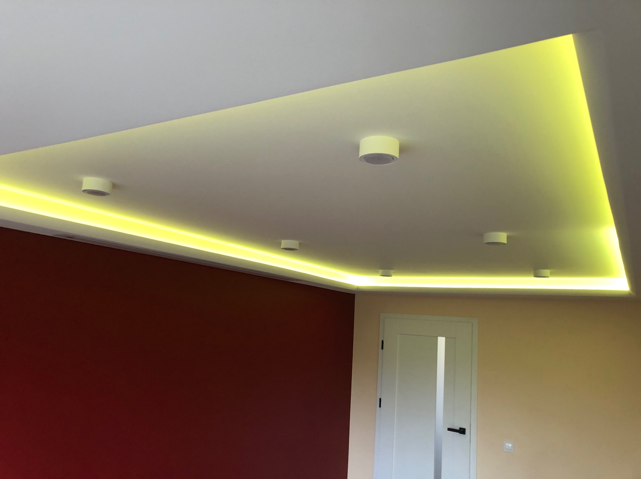 Podwieszany sufit z oświetleniem LED wzdłuż krawędzi, uzupełniony okrągłymi lampami sufitowymi, na tle ścian w odcieniach bordo i beżu oraz białych drzwi.
