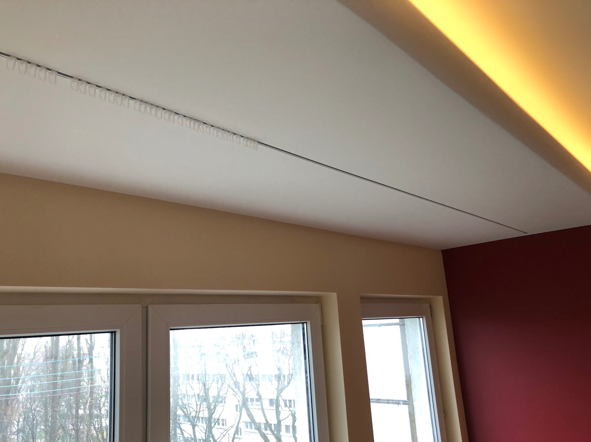 Biały sufit z zamontowaną szyną karniszową z haczykami, beżowe ściany i fragment ściany w kolorze burgundowym, widoczne okna z widokiem na drzewa.