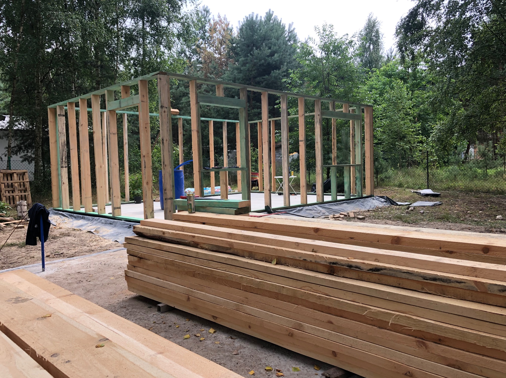 Konstrukcja szkieletowa budynku w trakcie budowy, widoczne drewniane belki i słupy, stos drewna na pierwszym planie, teren budowy zalesiony w tle.