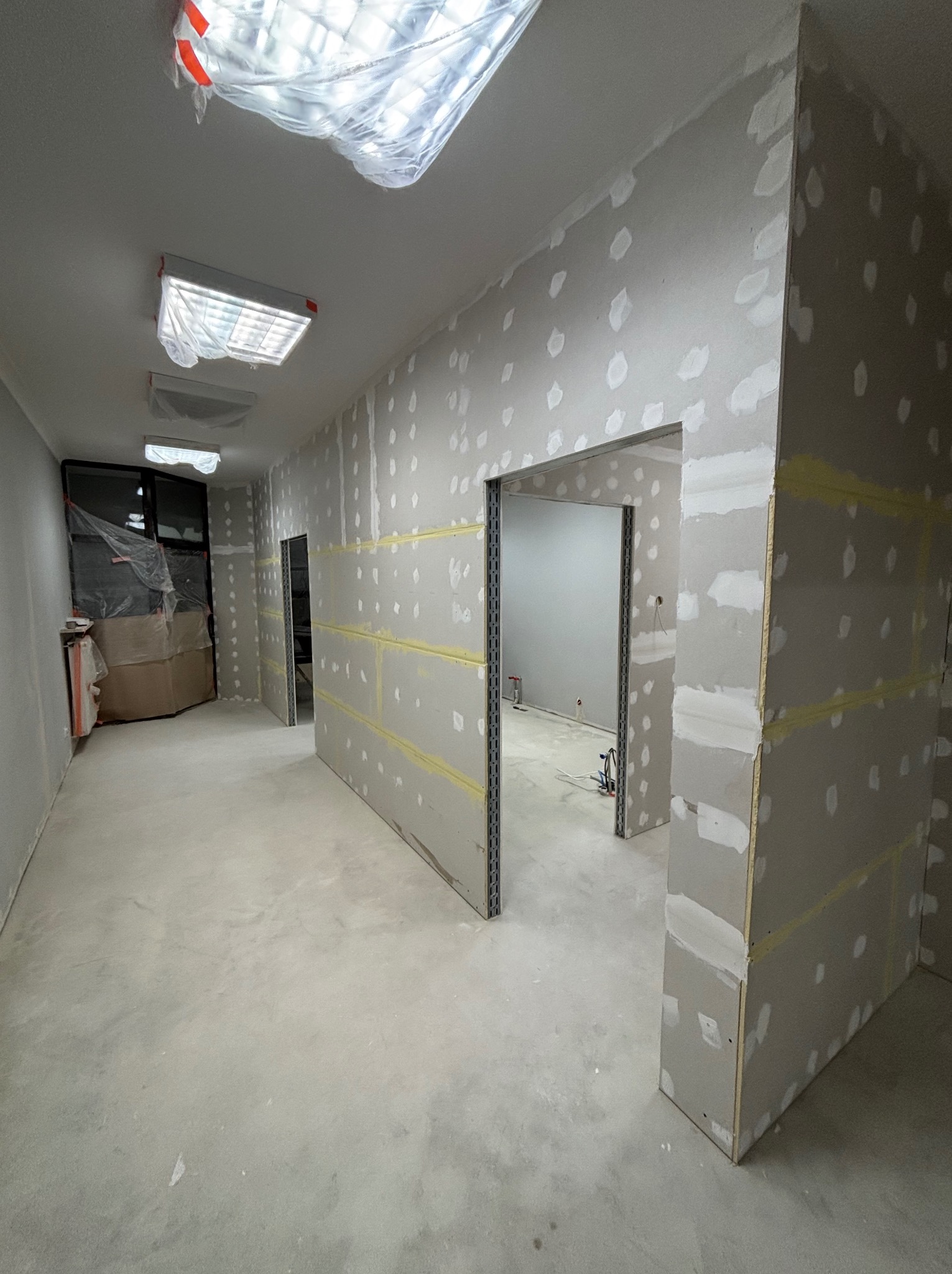 Wnętrze w trakcie prac wykończeniowych, widoczne płyty kartonowo-gipsowe z widocznymi punktami szpachlowania, metalowe profile ościeżnic, zabezpieczone folią lampy sufitowe.
