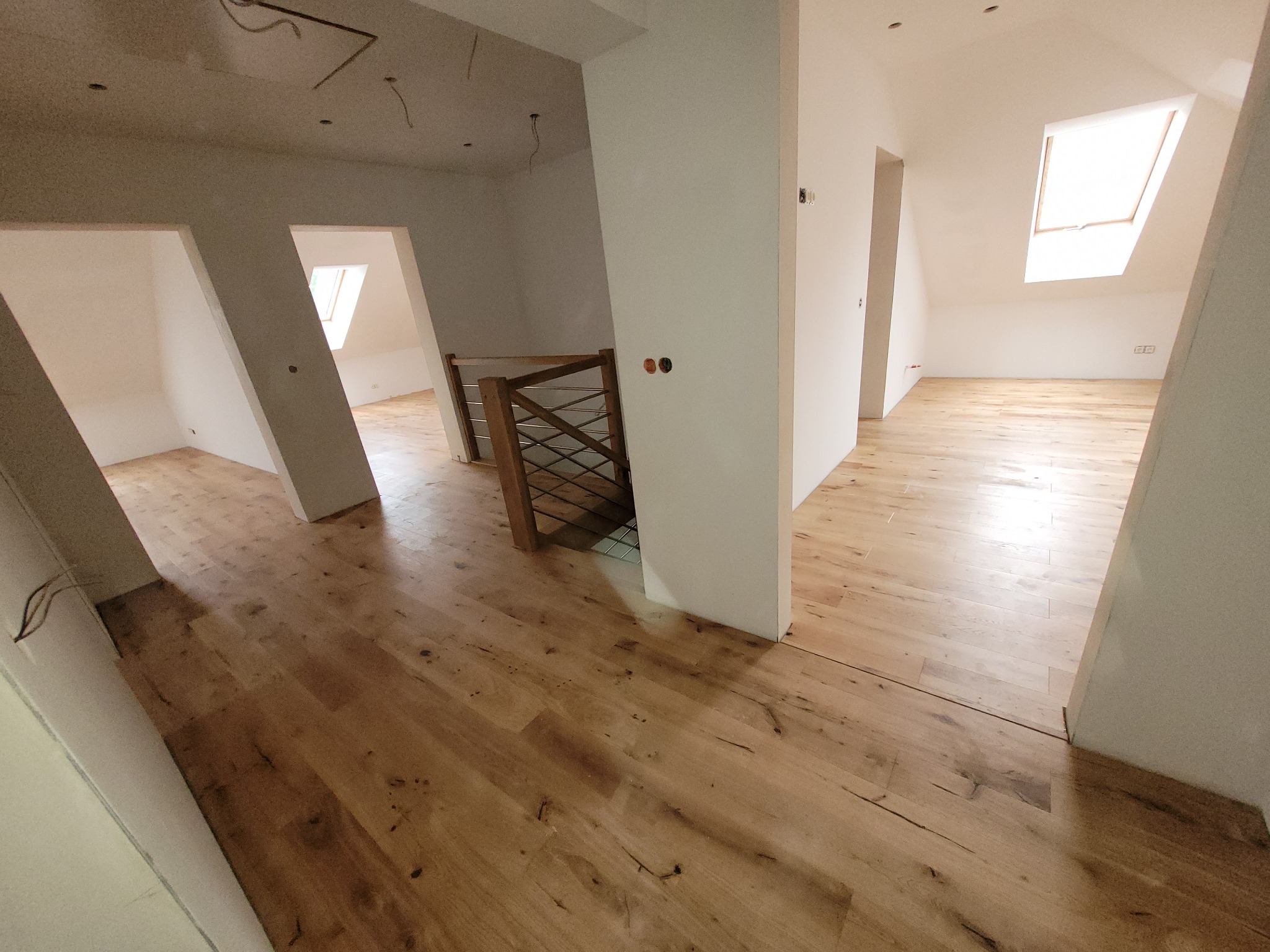 Jasna, drewniana podłoga ułożona w przestronnym, ale jeszcze surowym wnętrzu poddasza z widocznymi elementami konstrukcyjnymi i schodami z metalowymi barierkami.