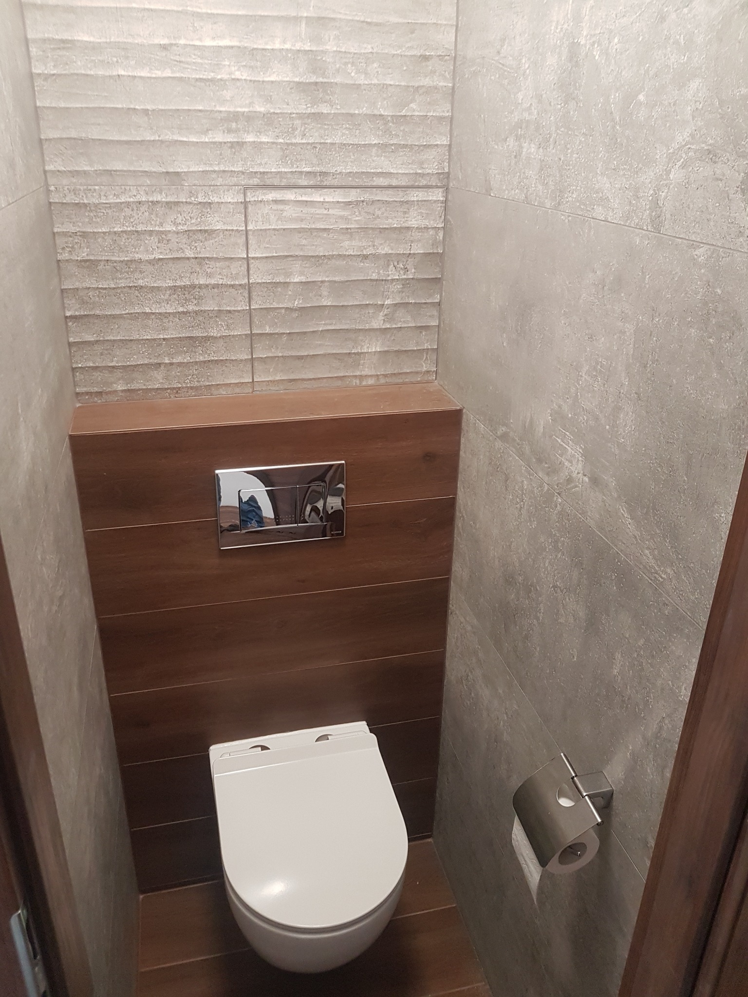 Nowoczesna, wykończona łazienka z podwieszaną toaletą, brązowymi i szarymi płytkami. Widoczny uchwyt na papier toaletowy i panel spłukujący.