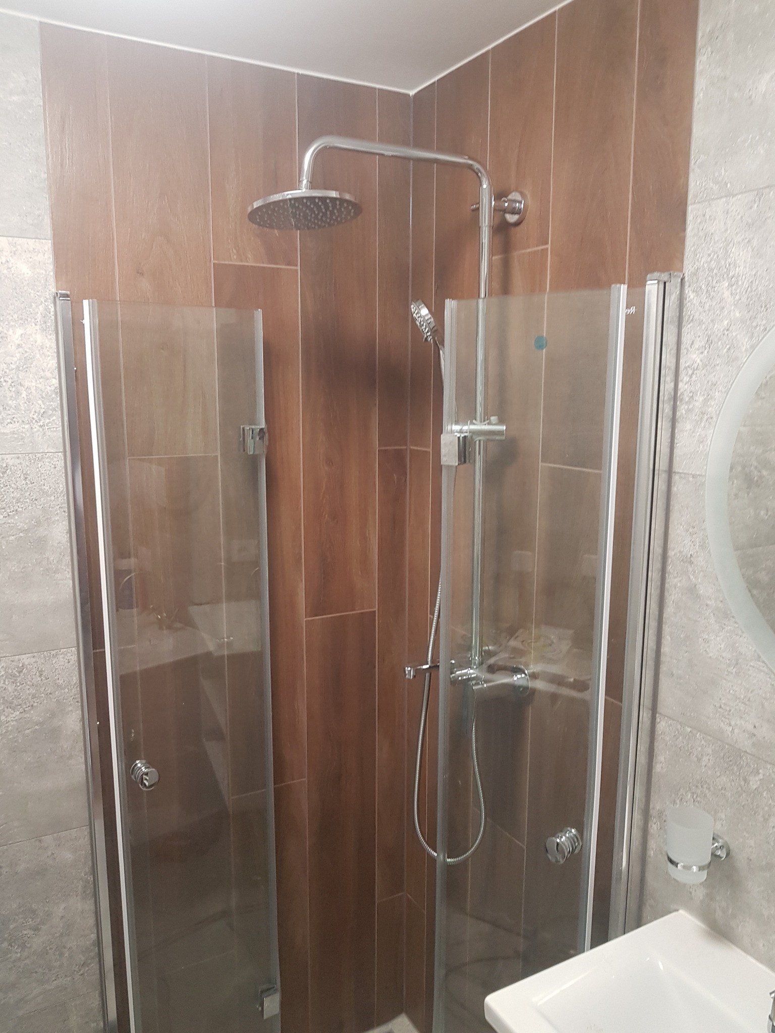 Nowoczesna kabina prysznicowa z deszczownicą i szklanymi drzwiami, wykończona płytkami imitującymi drewno. Widoczna umywalka i minimalistyczne akcesoria łazienkowe.