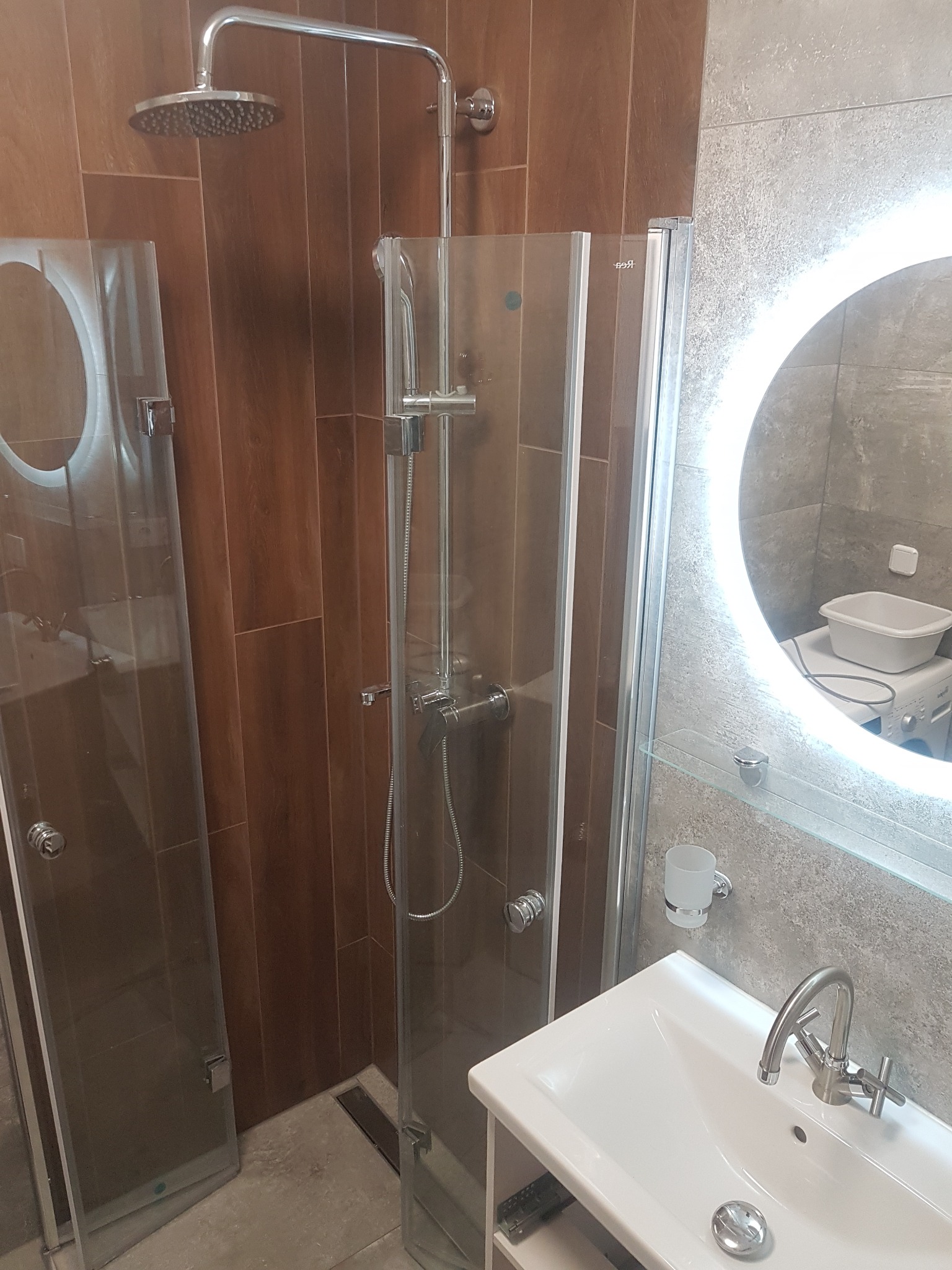 Nowoczesna łazienka z kabiną prysznicową typu walk-in, umywalką i okrągłym lustrem z podświetleniem LED. Brązowe płytki imitujące drewno na ścianie.