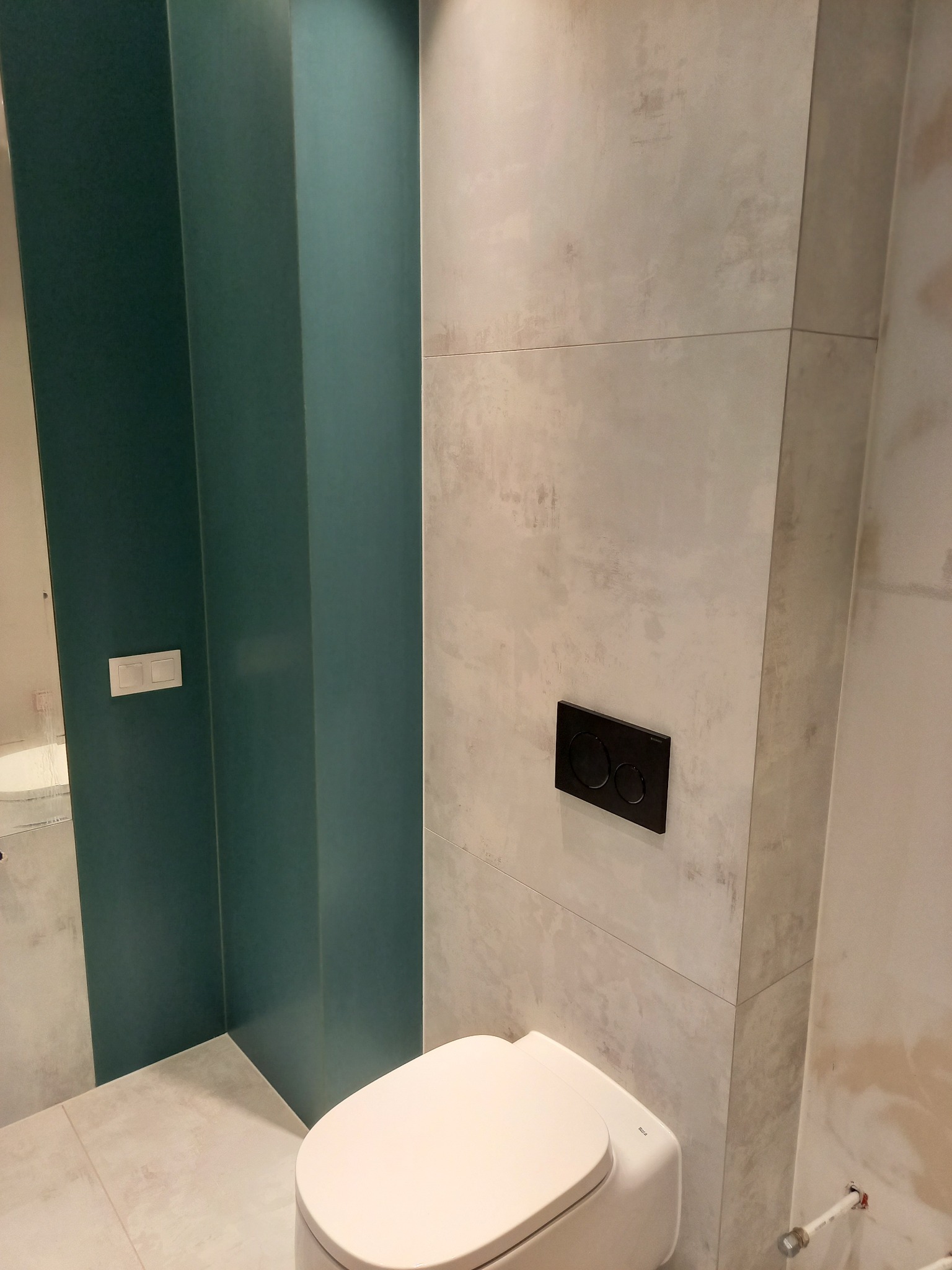 Nowoczesna toaleta podwieszana z czarnym panelem spłukującym na tle płytek imitujących beton i zielonej zabudowy. Minimalistyczny design w wykończonej łazience.