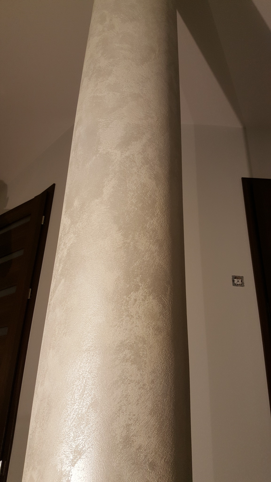 Dekoracyjny filar w jasnych odcieniach beżu z efektem strukturalnym, widoczny fragment ściany i drzwi z ciemnego drewna. Całość w ciepłym oświetleniu.