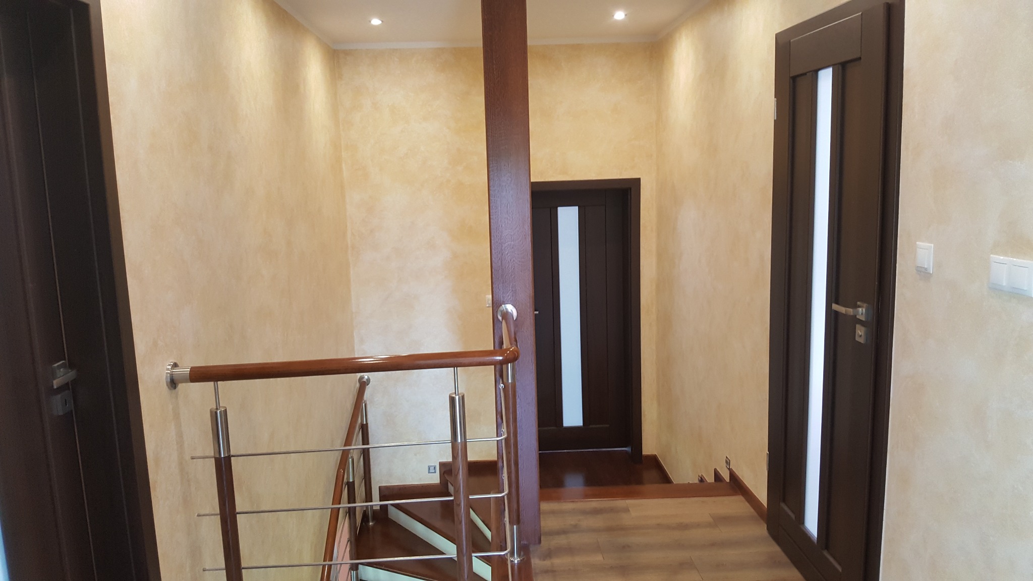Klatka schodowa z wykończeniem ścian Glinką Wenecką w ciepłych tonacjach. Widoczne drewniane schody, brązowe drzwi i metalowa balustrada.