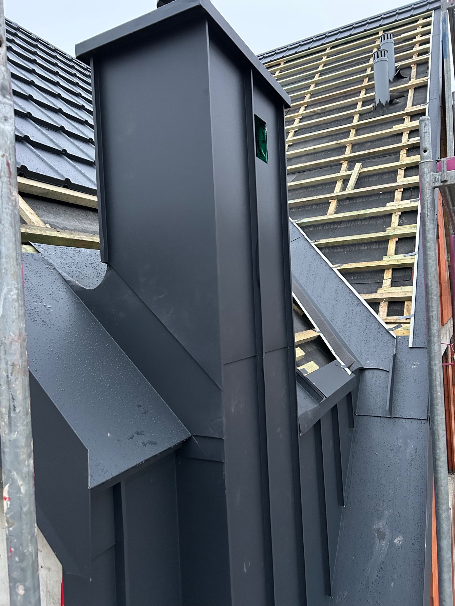 Nowoczesny, grafitowy komin z blachy na dachu z dachówką ceramiczną; w tle widoczna konstrukcja więźby dachowej z łatami i kontrłatami, obróbki blacharskie wykończone z dbałością o detale.