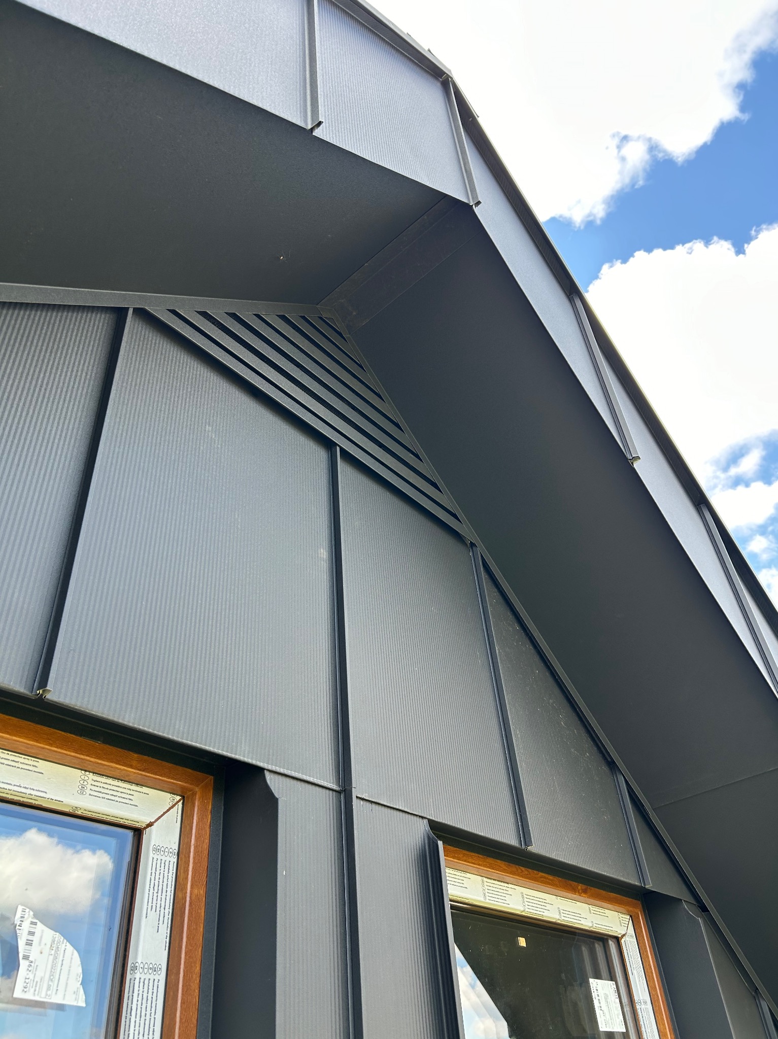 Grafitowa obróbka blacharska na budynku z widocznymi oknami i fragmentem nieba z chmurami.