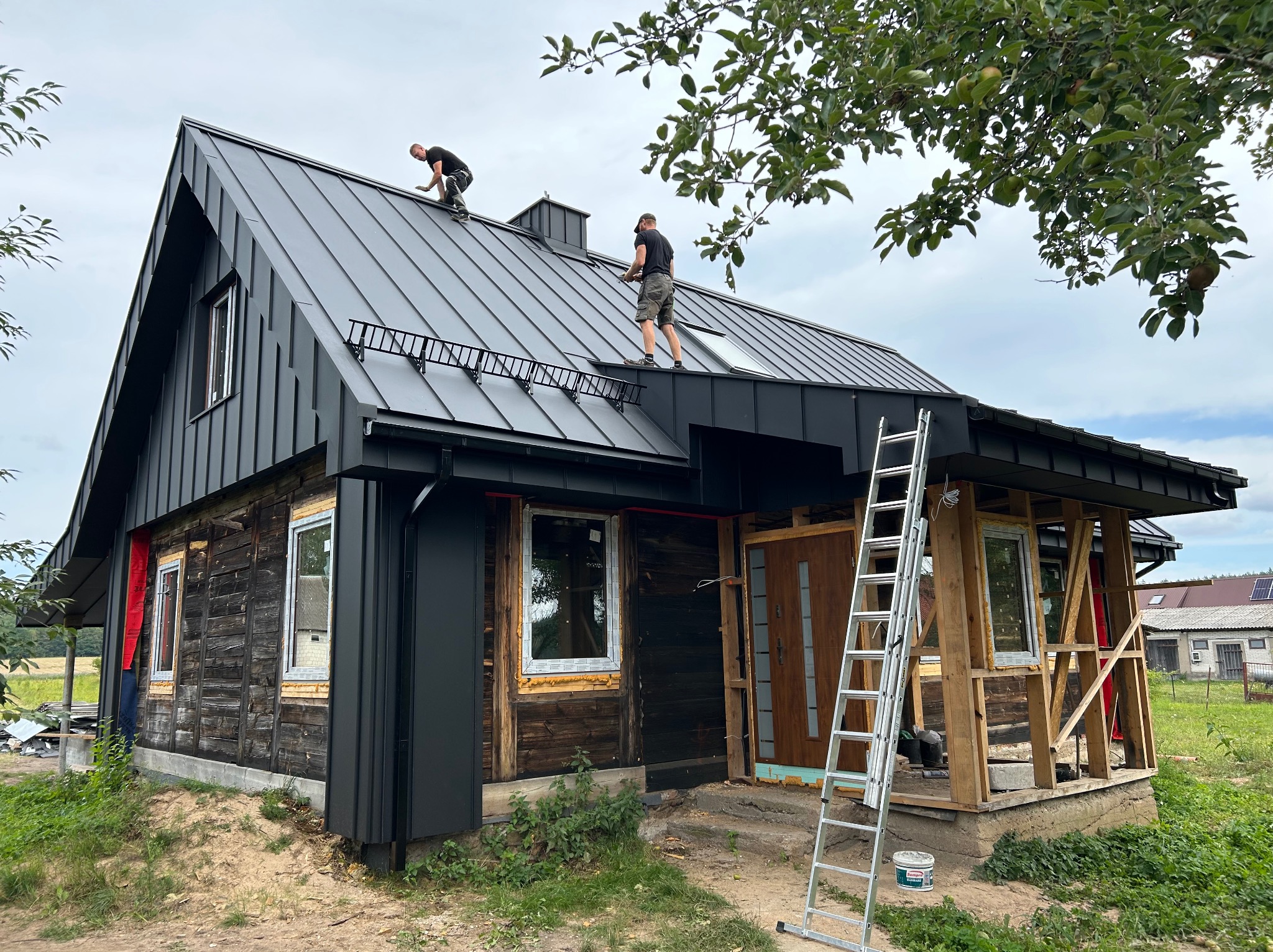 Dwóch dekarzy pracuje na dachu domu z ciemnej blachy, widoczna drabina i częściowo wykończona elewacja z drewnianymi elementami oraz fragmenty drzewa owocowego z jabłkami.